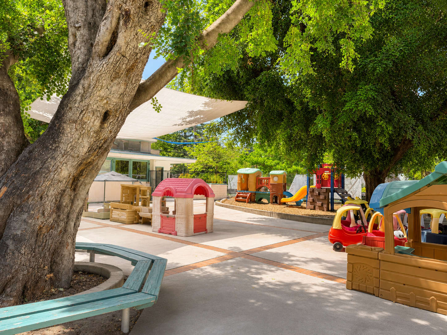 Children's playground with colorful plastic playhouses, slides, and sandbox under large leafy trees, with shaded seating area. WHCCNS