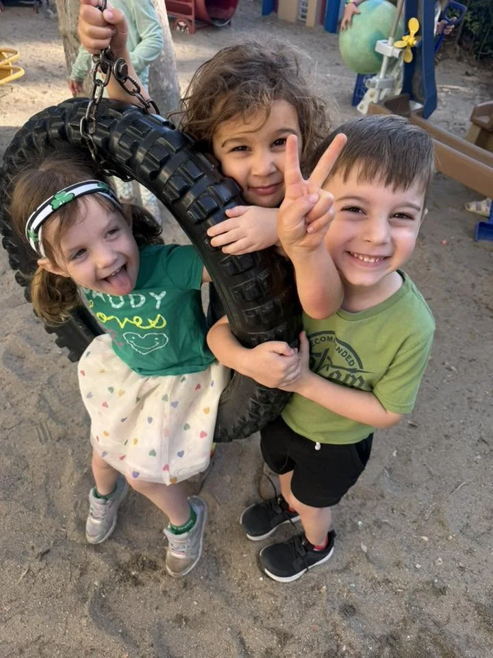 Three children playing on a tire swing at a playground, smiling and making peace signs. WHCCNS