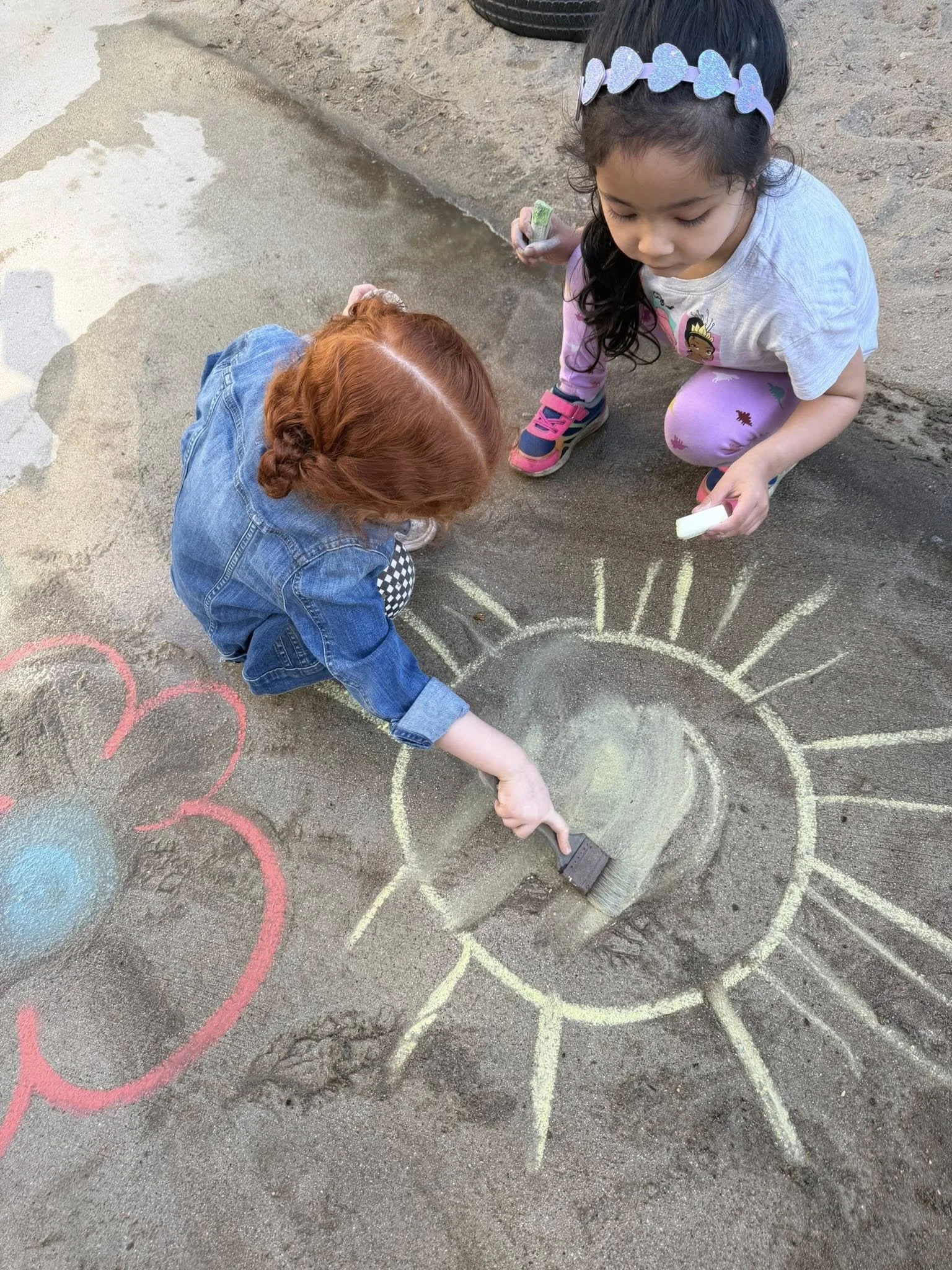 Two young girls drawing with chalk on the ground, creating a rainbow.