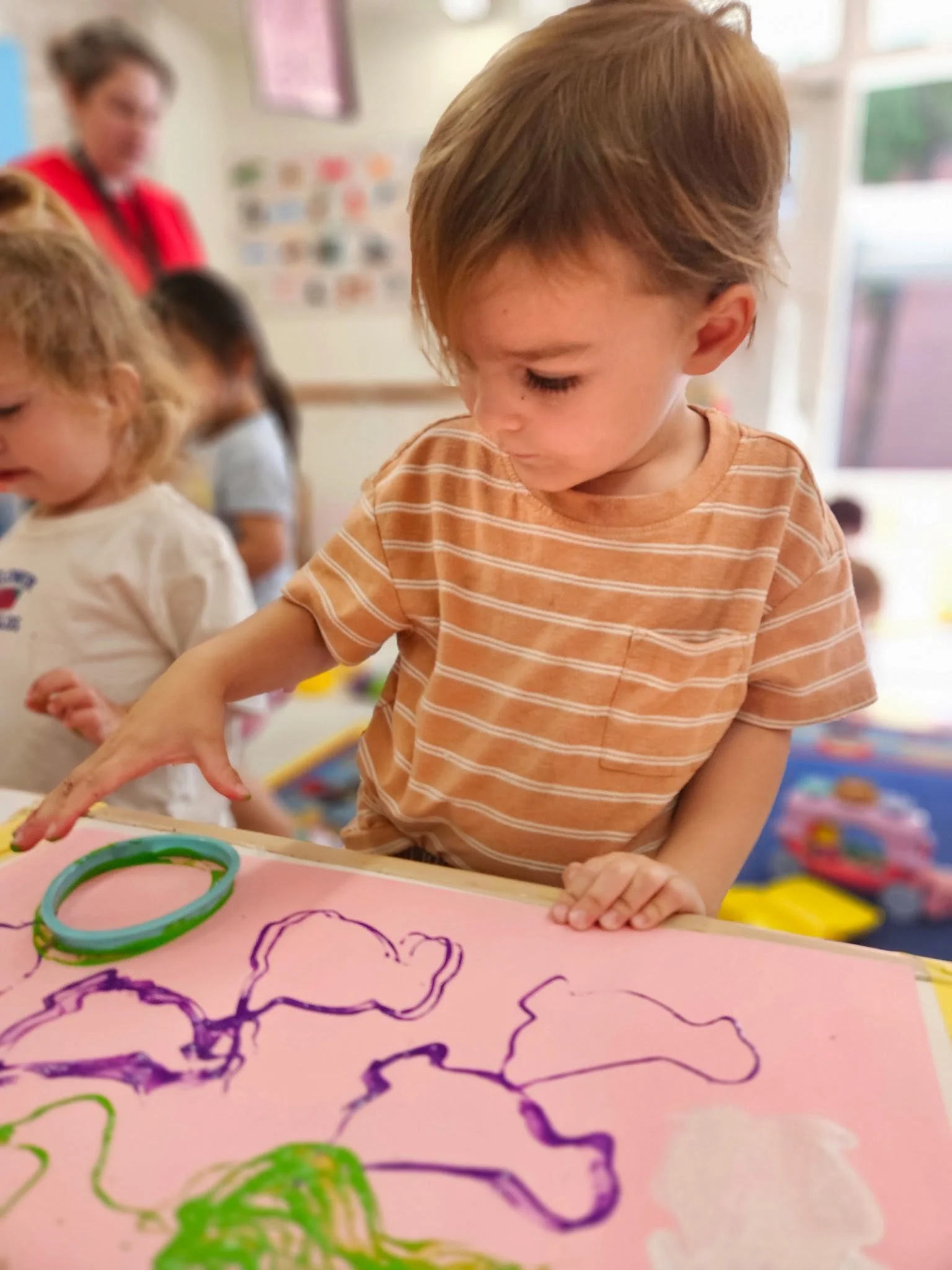 A young boy in a peach and white striped shirt is drawing with purple, green, and black markers on pink paper at a table. Other children and a teacher are in the background at WHCCNS