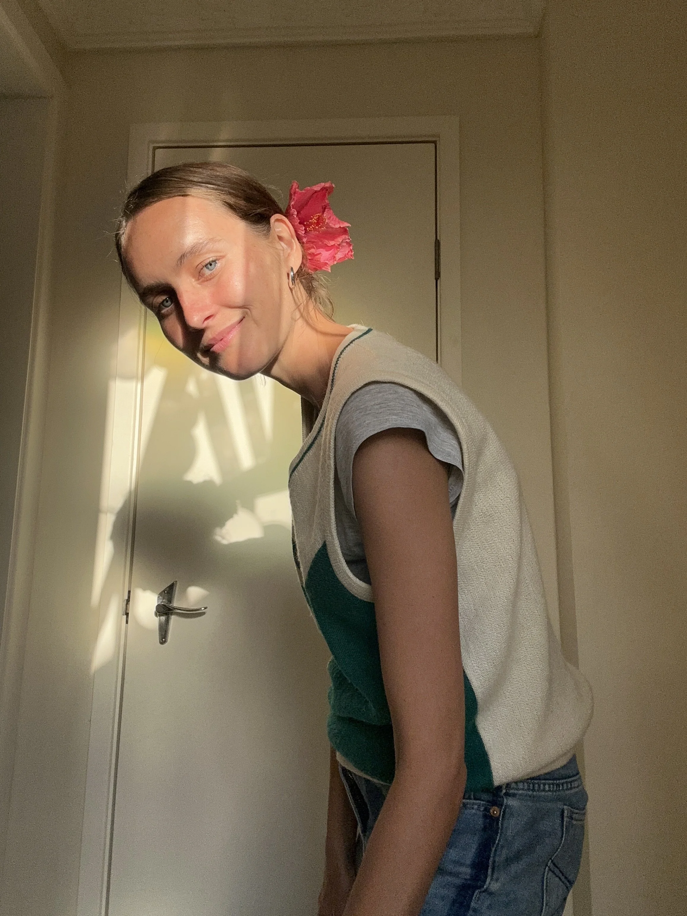 A woman with light-colored eyes and brown hair tied back in a ponytail, wearing a sleeping mask or eye mask on her forehead, a sleeveless shirt with colored panels, and blue jeans, standing in a room near a door with a pink flower behind her ear.