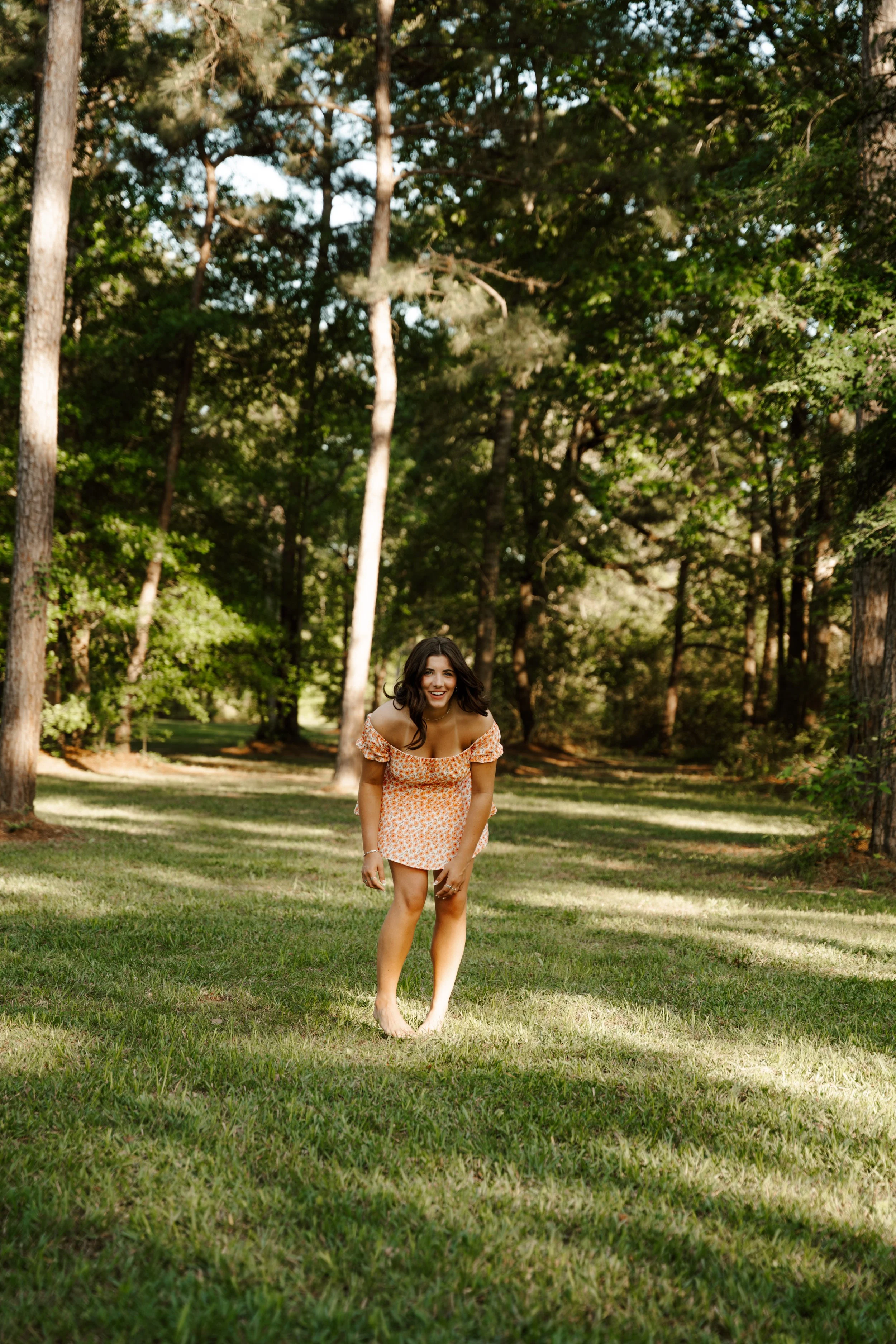 A young woman with dark hair is smiling and bending forward slightly while standing barefoot on the grass in a wooded park.