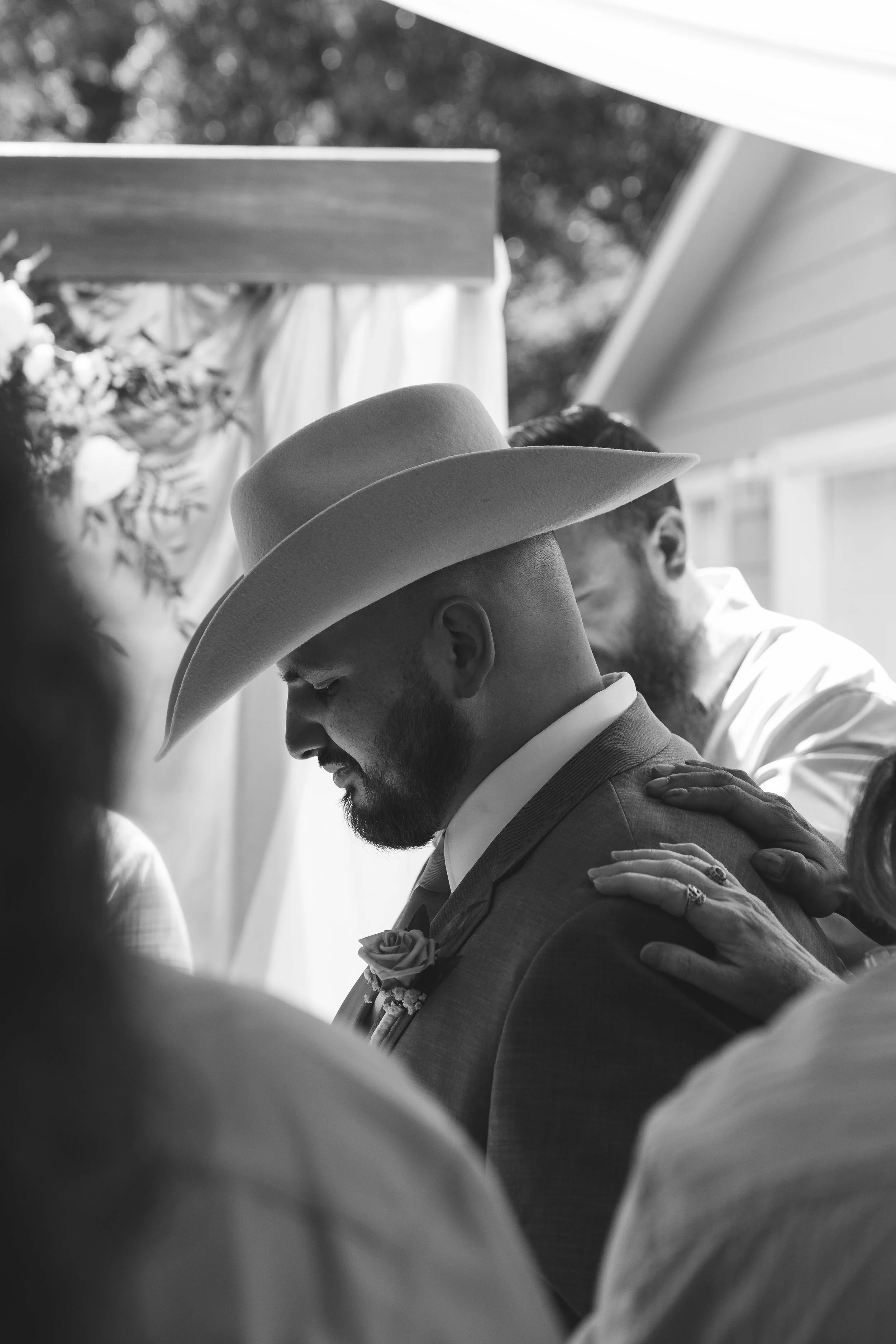 Black and white photograph of a man in a suit and wide-brimmed hat with a beard and mustache, with his head bowed and eyes closed, and a woman with her hand on his shoulder, in what appears to be a prayer or contemplative moment, with other people pa
