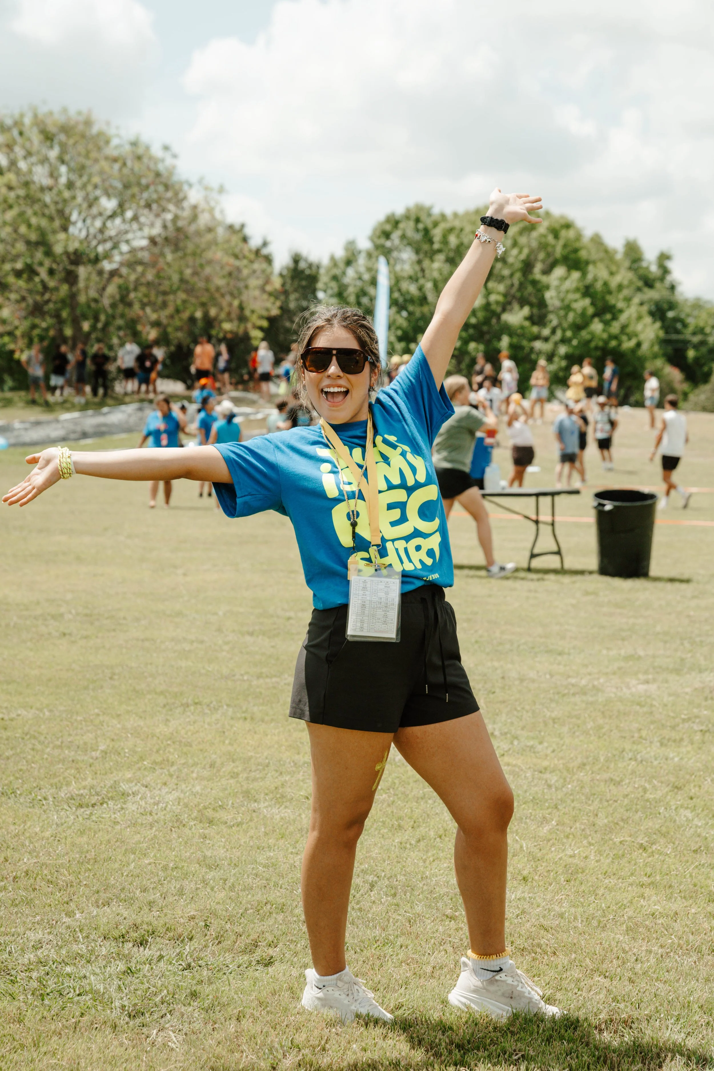 A woman wearing sunglasses, a blue T-shirt with yellow text, black shorts, and white sneakers standing outdoors on a grassy field with her arms outstretched, smiling with her mouth open. In the background, there are many people, trees, and cloudy ski