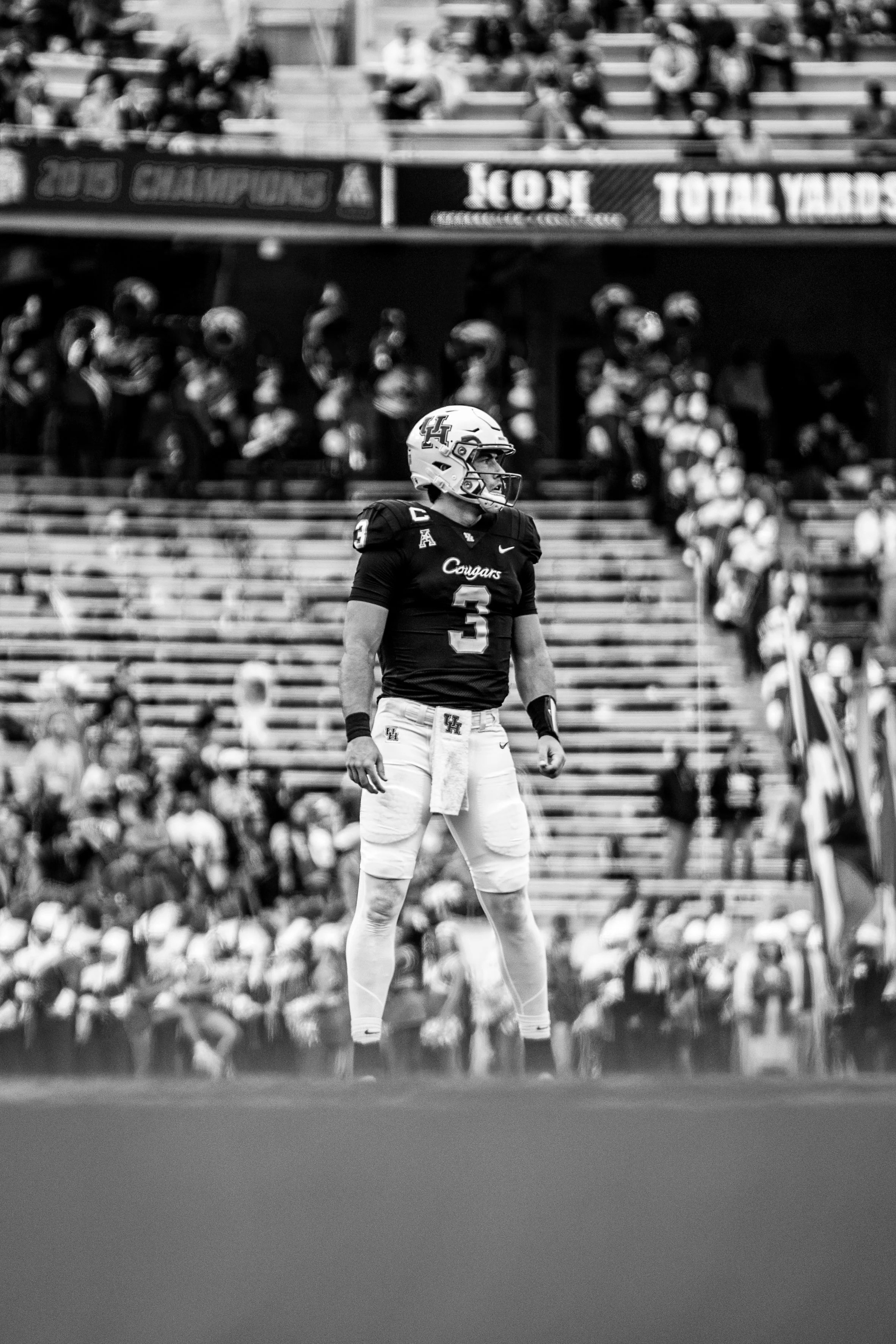 A football player wearing a Houston Cougars uniform stands on the field during a game, with spectators in the stadium stands behind him.