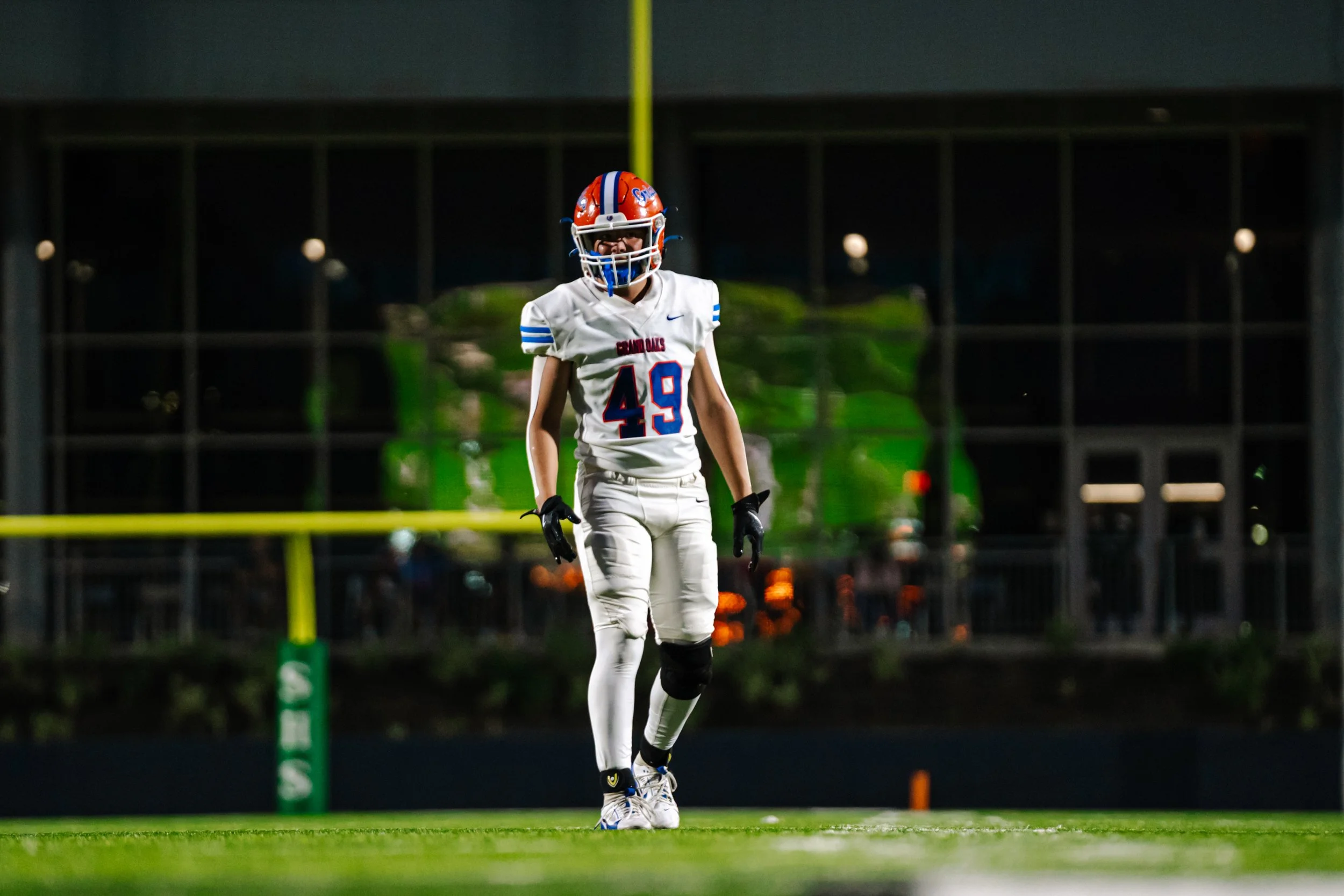 A football player wearing a white uniform with blue and red accents, number 49, and a red helmet, standing on a football field at night, with a blurred background and visible goalpost.