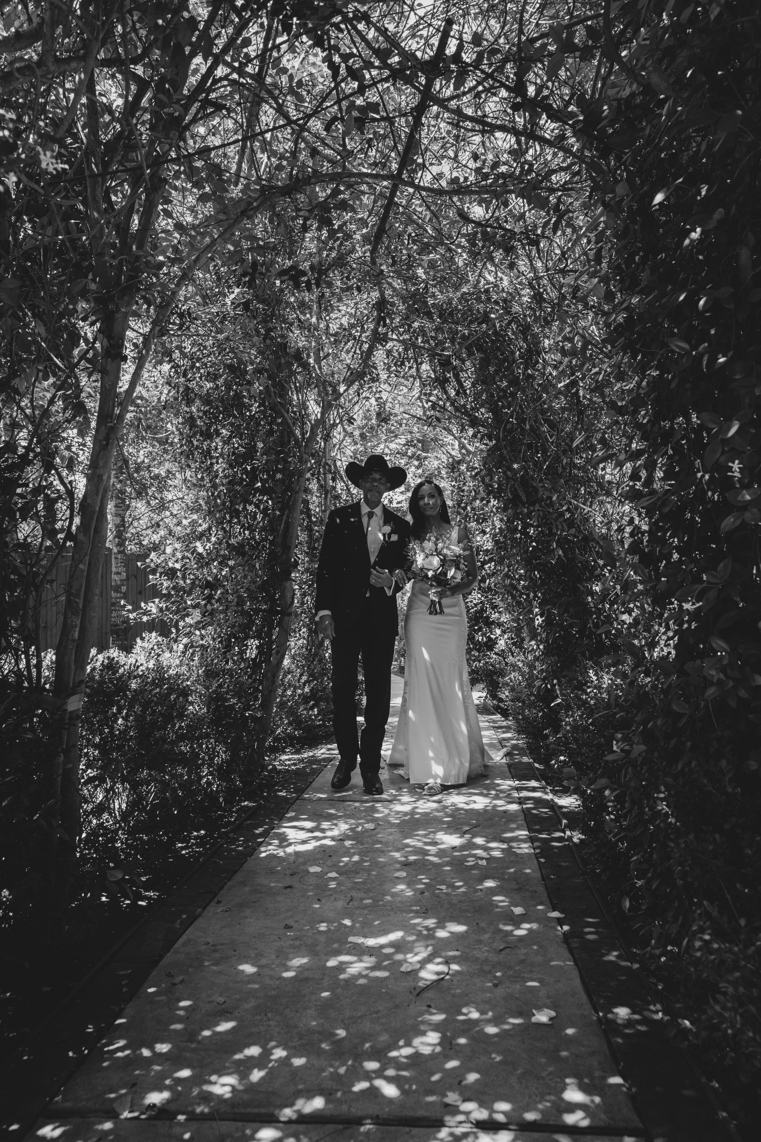 A black and white photograph of a wedding couple walking down a garden pathway surrounded by leafy trees and bushes. The groom is wearing a dark suit and cowboy hat, and the bride is wearing a white gown holding a bouquet of flowers.