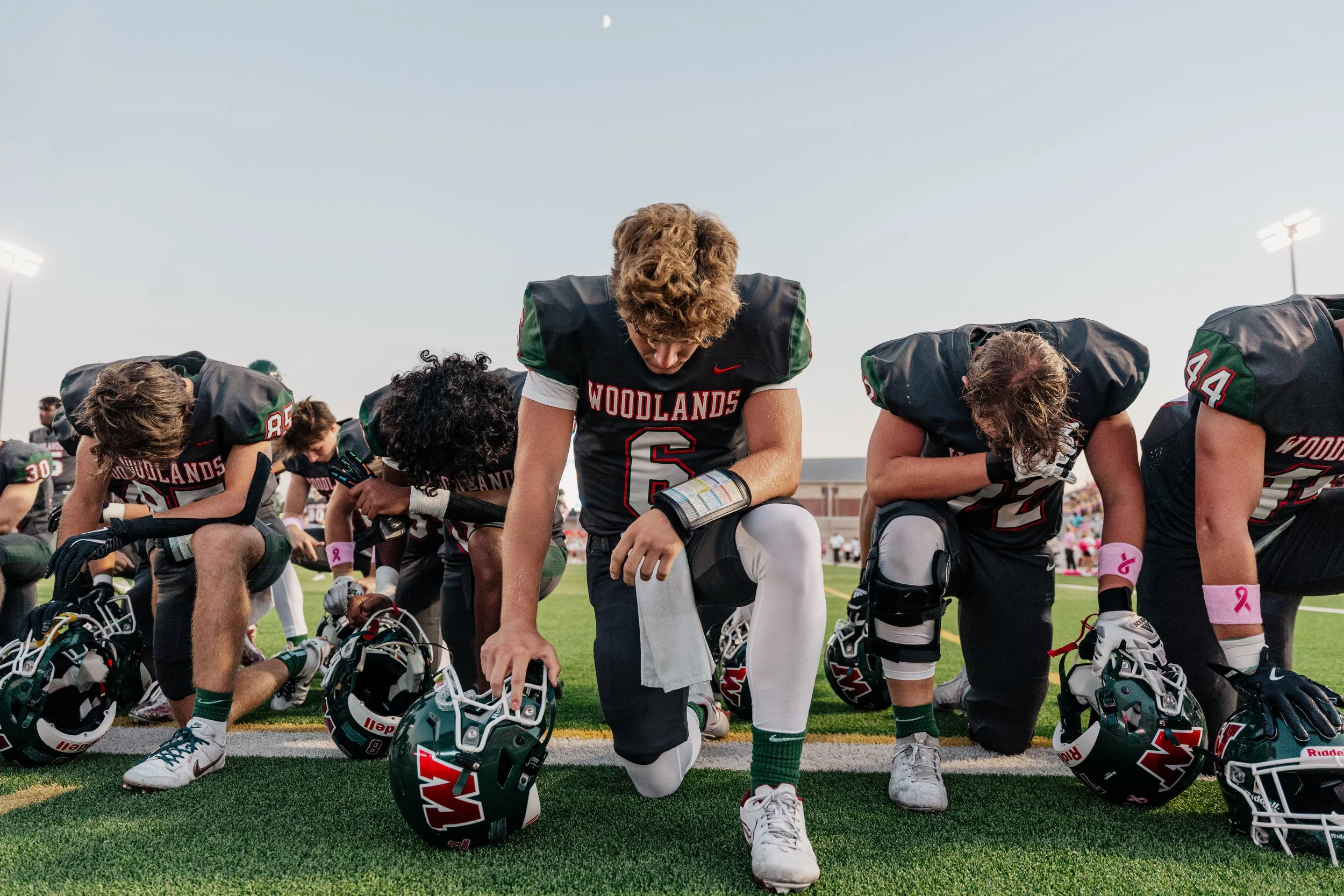American football players kneeling on the field during a pre-game prayer, wearing black jerseys with red and green accents, holding their helmets, with a stadium and night sky in the background.