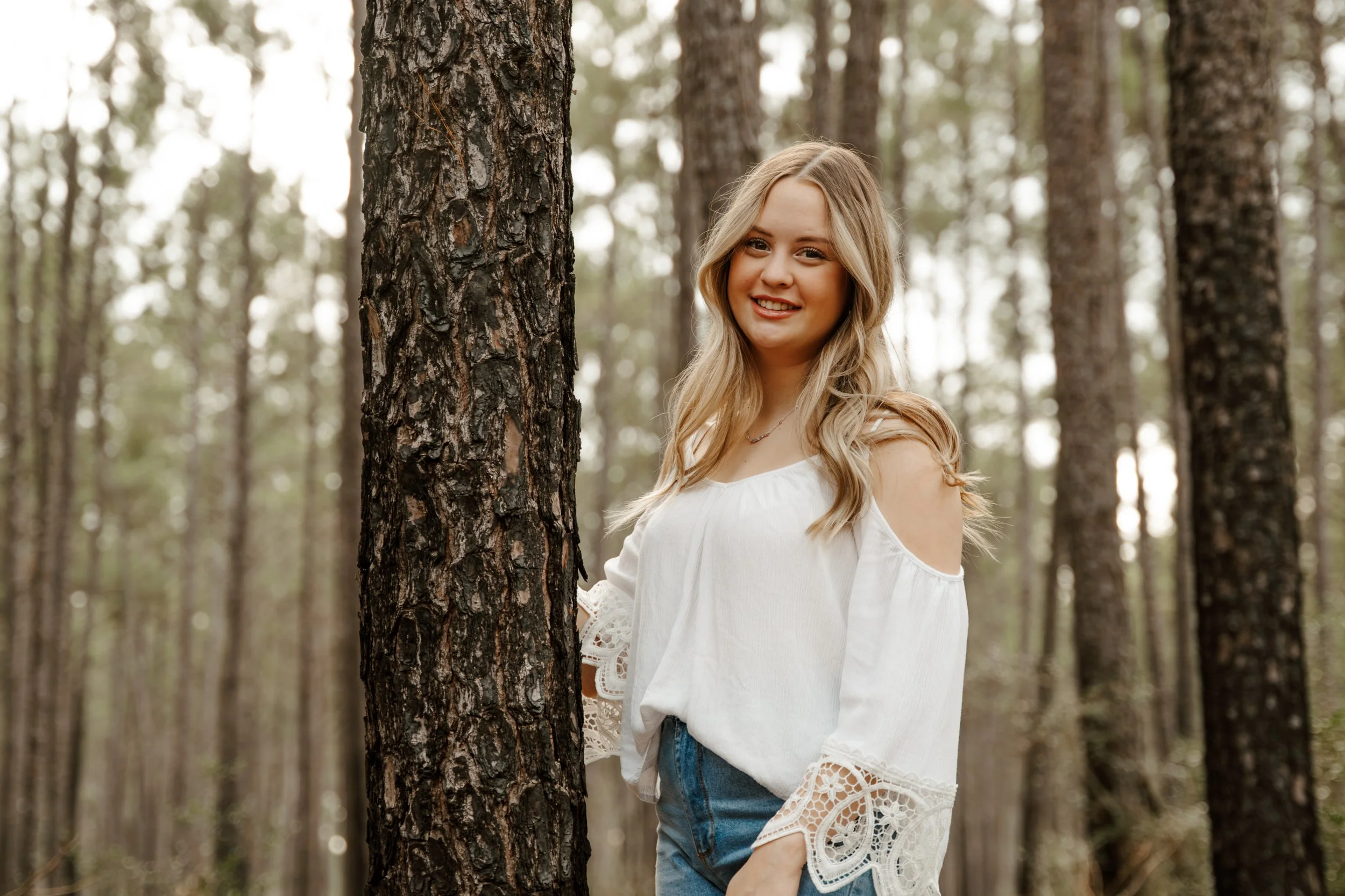 Young woman with long blonde hair smiling in a white embroidered blouse in a forest with tall trees.