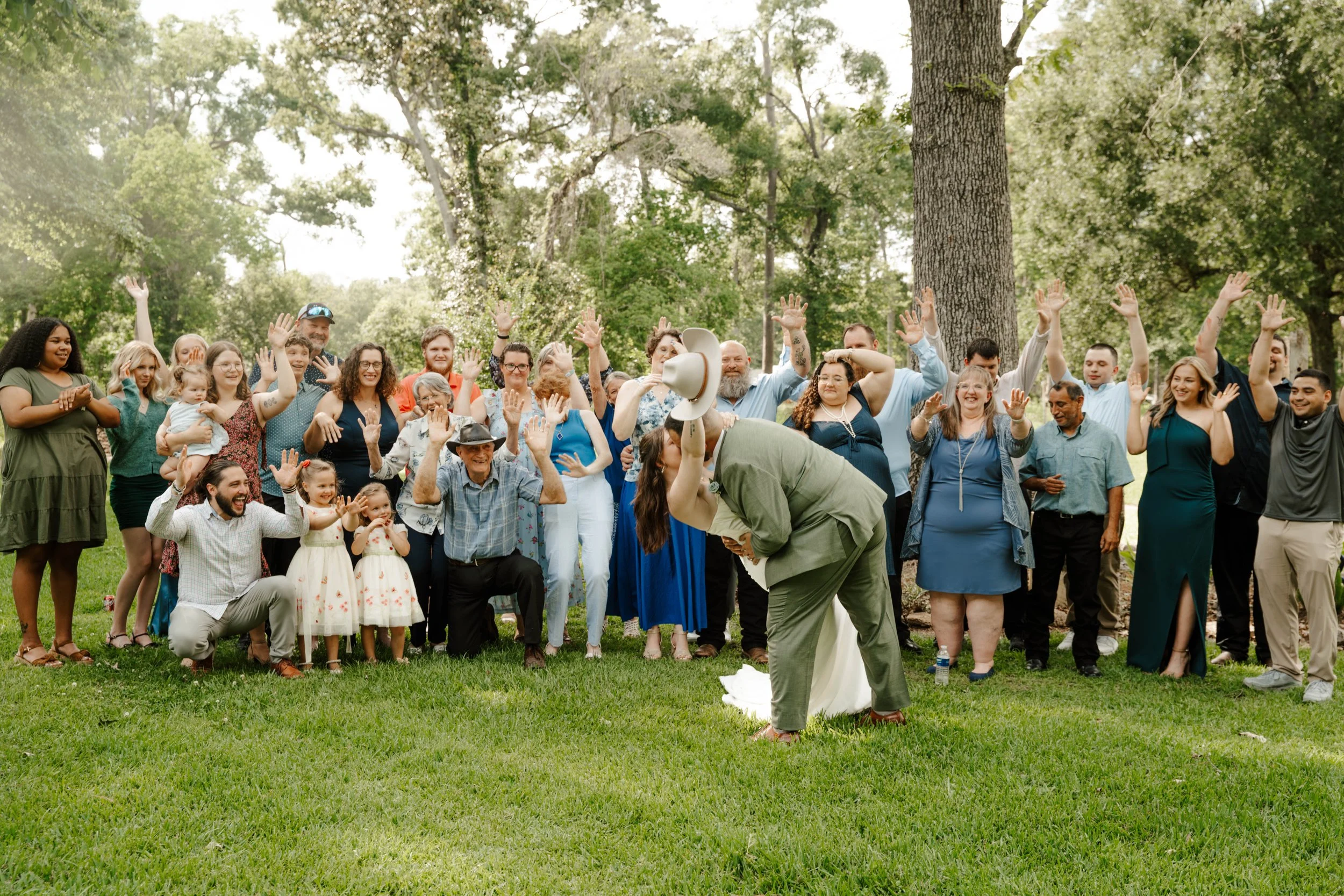 A large group of diverse people outdoors in a park, smiling and raising their hands for a group photo, with tall trees in the background.