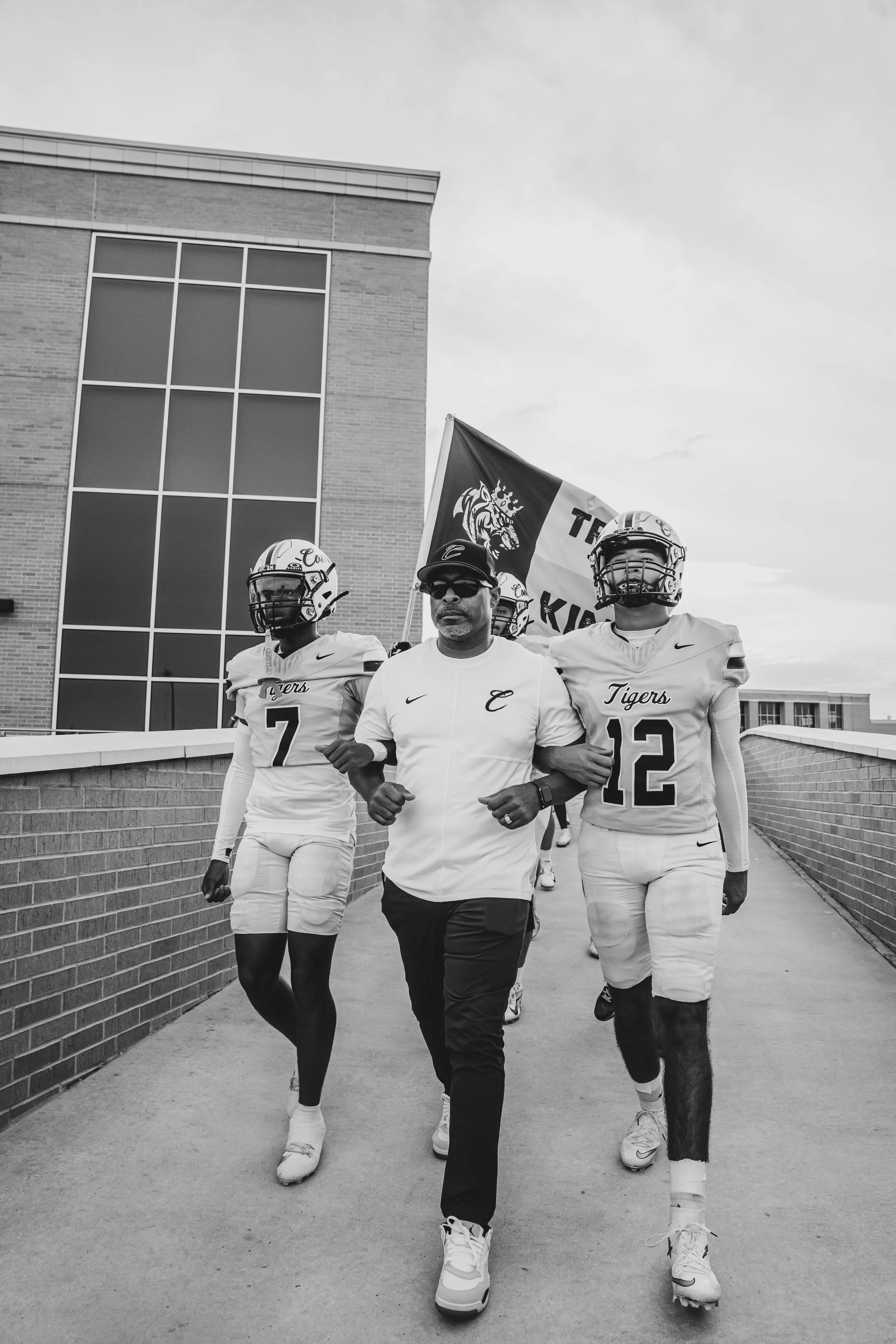 A group of football players and a coach walking together outside a building. The players are wearing helmets and football uniforms labeled "Tigers". The coach is in casual attire with sunglasses and a hat, holding a flag with a tiger and the words "T