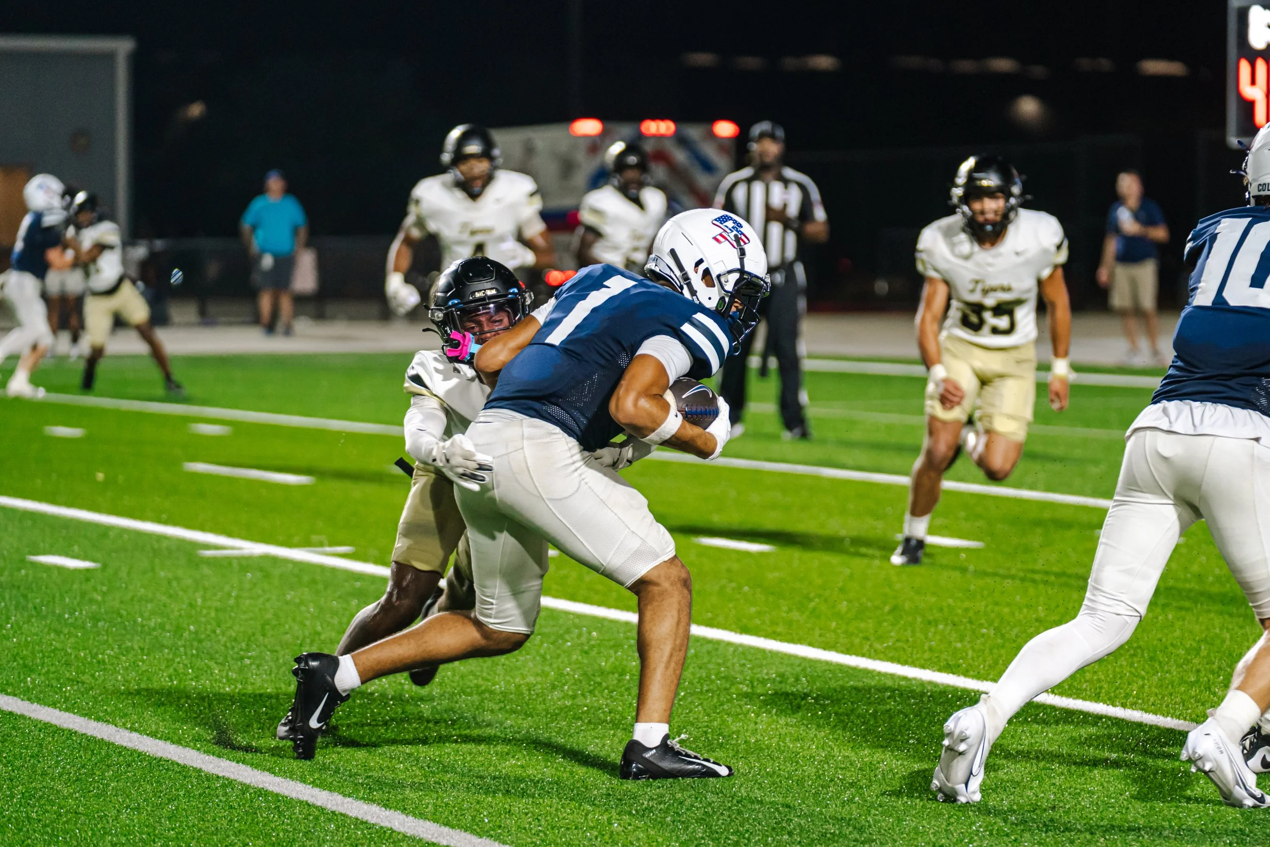 Young football player in white uniform and black helmet tackles a player in navy blue uniform with a white helmet holding a football on a brightly lit football field at night, with other players and officials in the background.