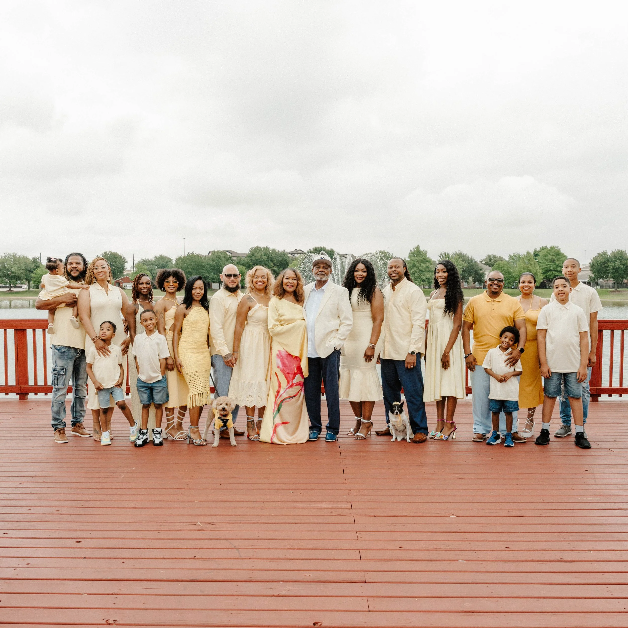 Group of people standing on a wooden dock near a lake, smiling and posing for a photo, some with children and dogs, in an outdoor park setting with trees and houses in the background.