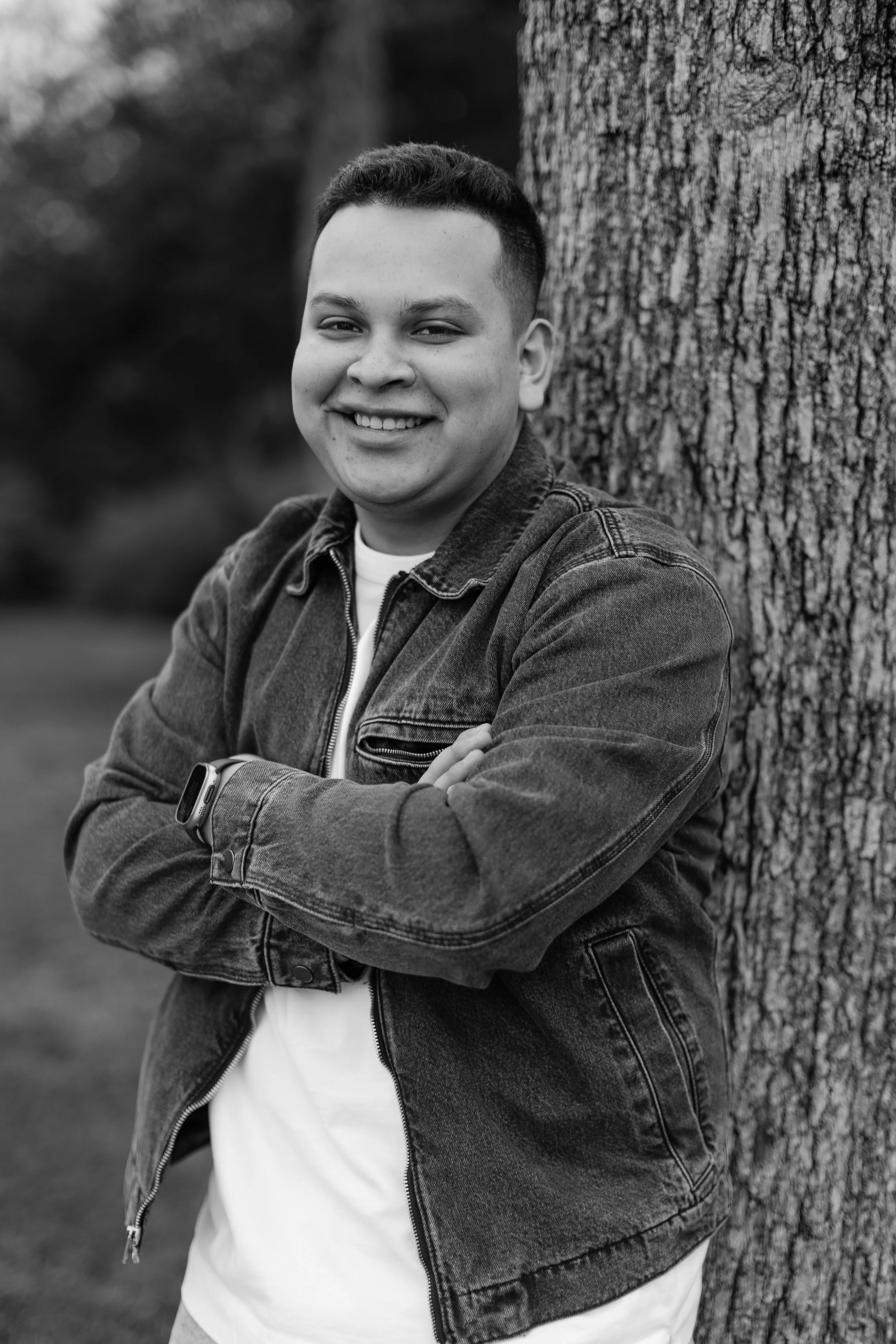 A young man with short dark hair smiling, leaning against a large tree trunk outdoors in a park or natural setting, wearing a denim jacket over a white t-shirt.