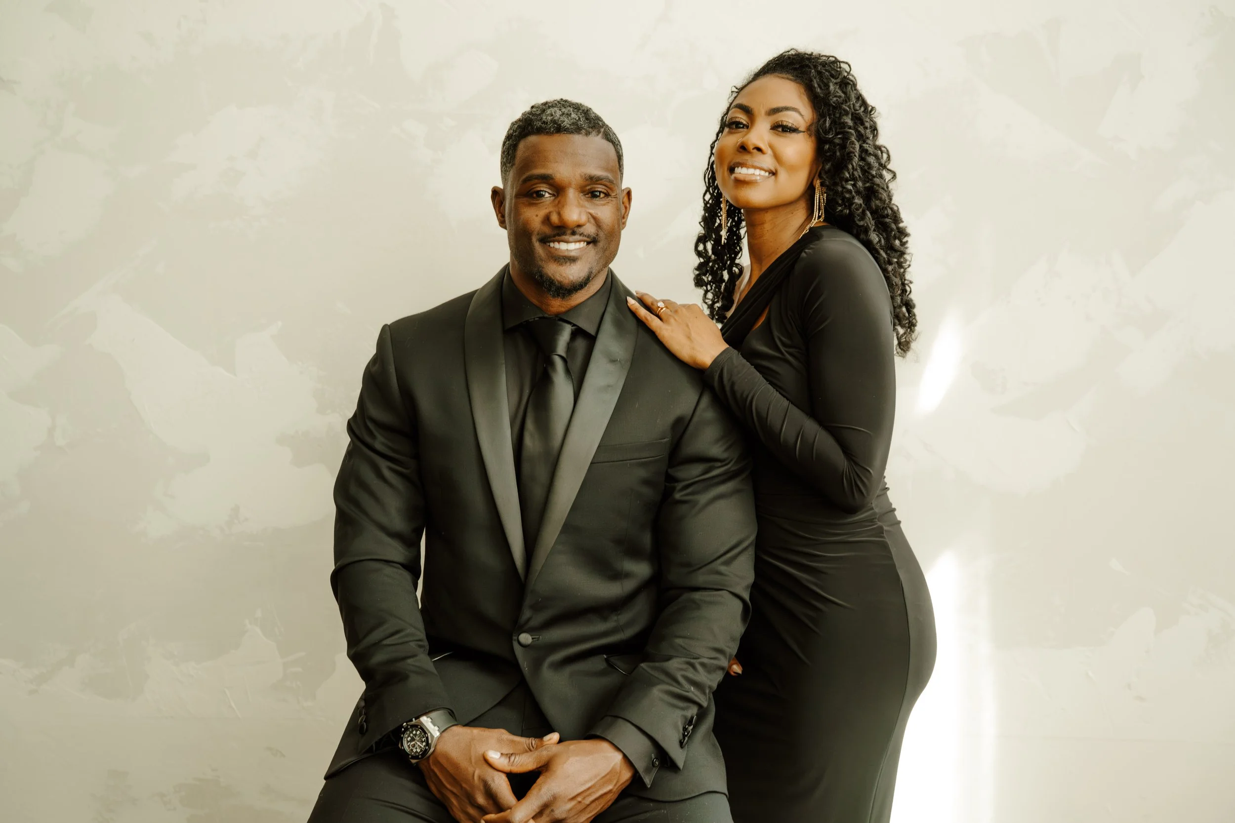 A man and woman dressed in black sitting close together, smiling at the camera, with the woman resting her hand on the man's shoulder.