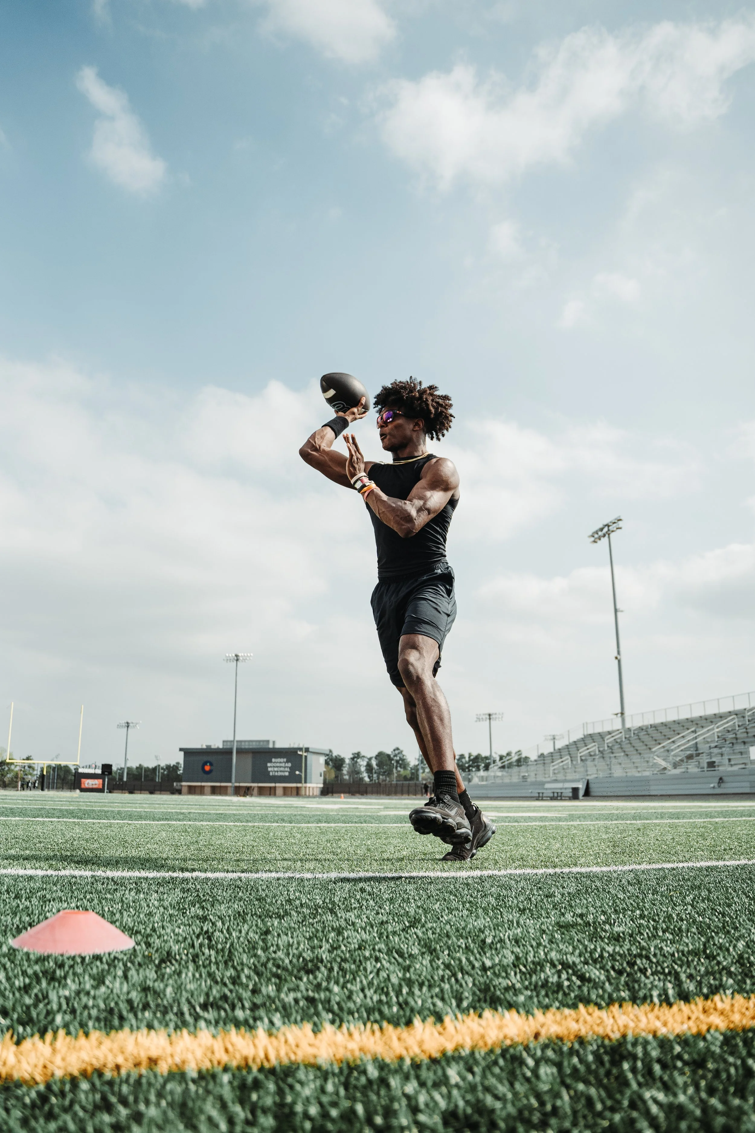A football player practicing on an outdoor football field, wearing sunglasses and running with a football in his right hand.