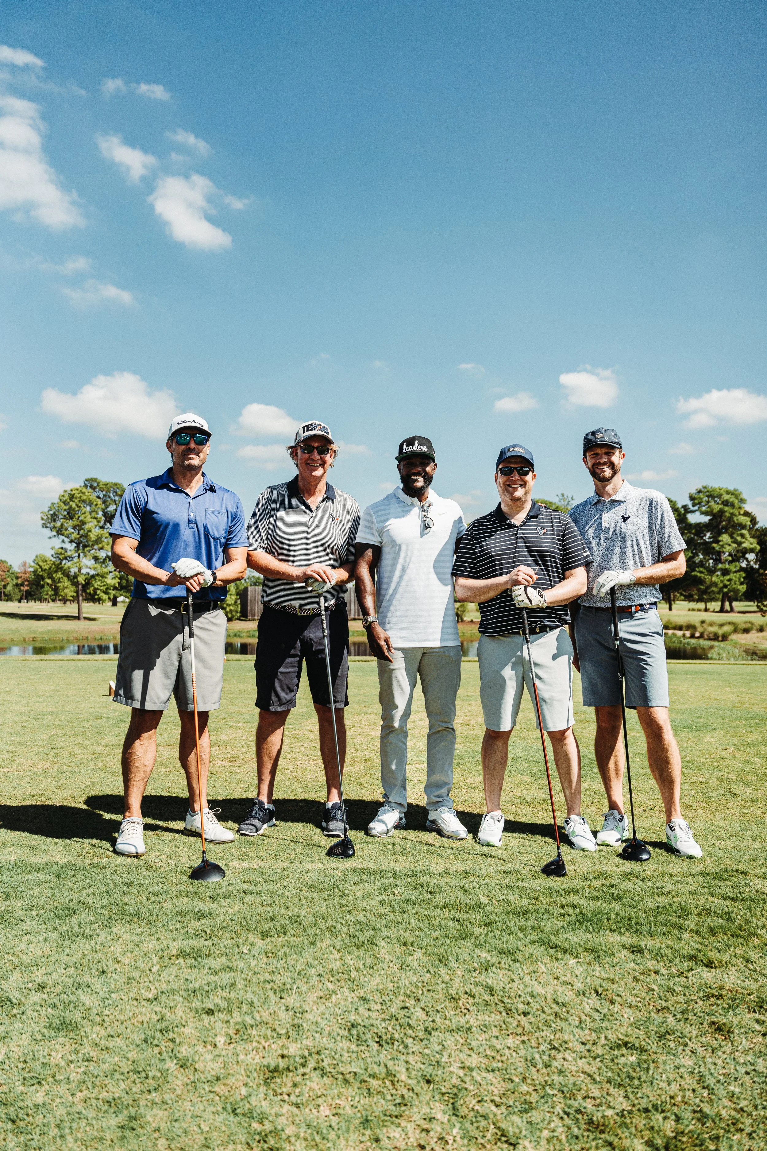 Five men standing on a golf course, smiling and holding golf clubs, wearing casual golf attire, with trees and a blue sky in the background.