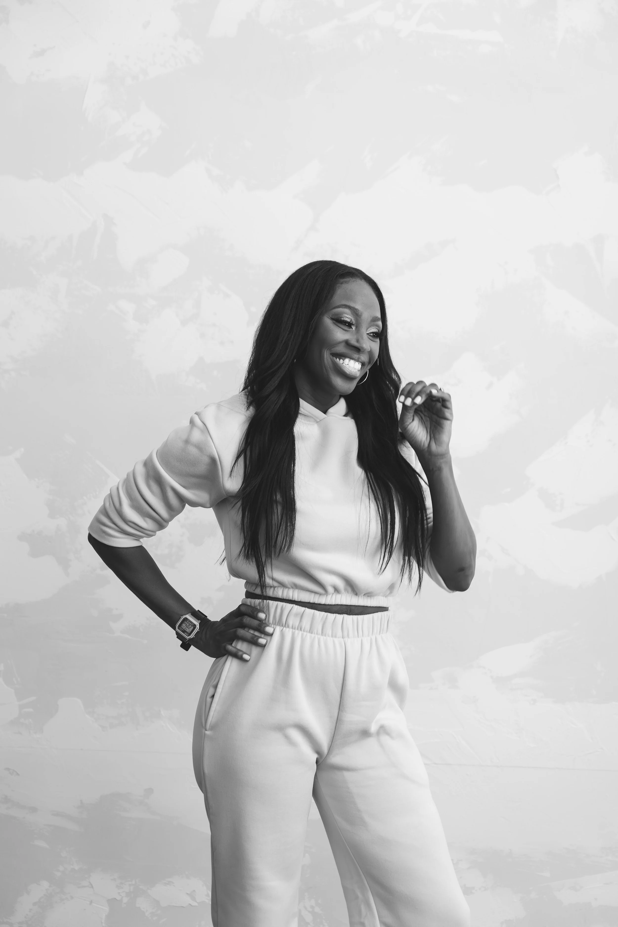 A woman smiling and posing with her left hand on her hip, wearing a light-colored tracksuit with a hoodie and high-waisted sweatpants, standing against an abstract background.