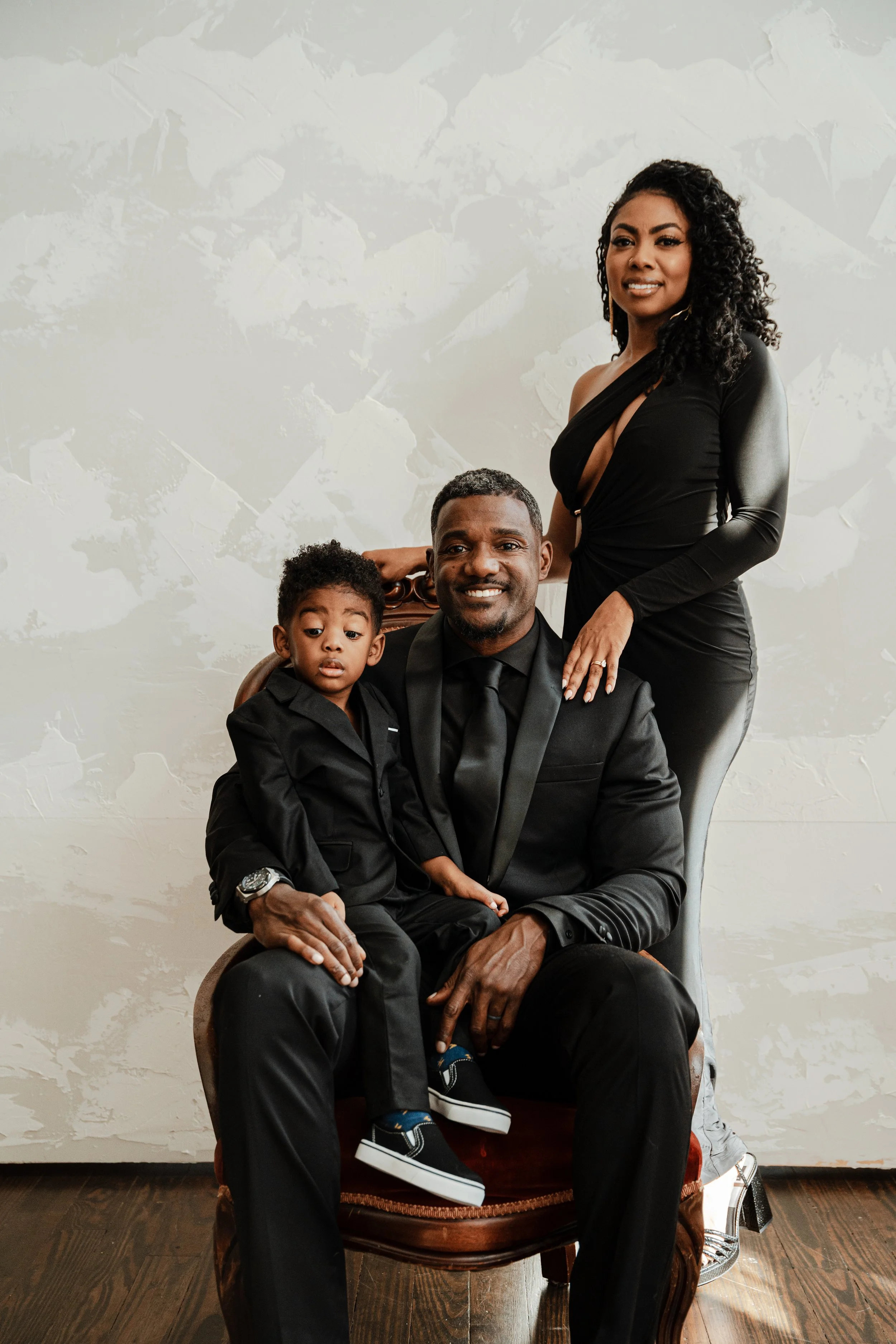A family portrait of an African American man, woman, and young boy. The man is sitting in a chair wearing a black suit, and the woman and boy are standing and sitting behind him, respectively, all dressed in black. The woman has curly hair and is smi