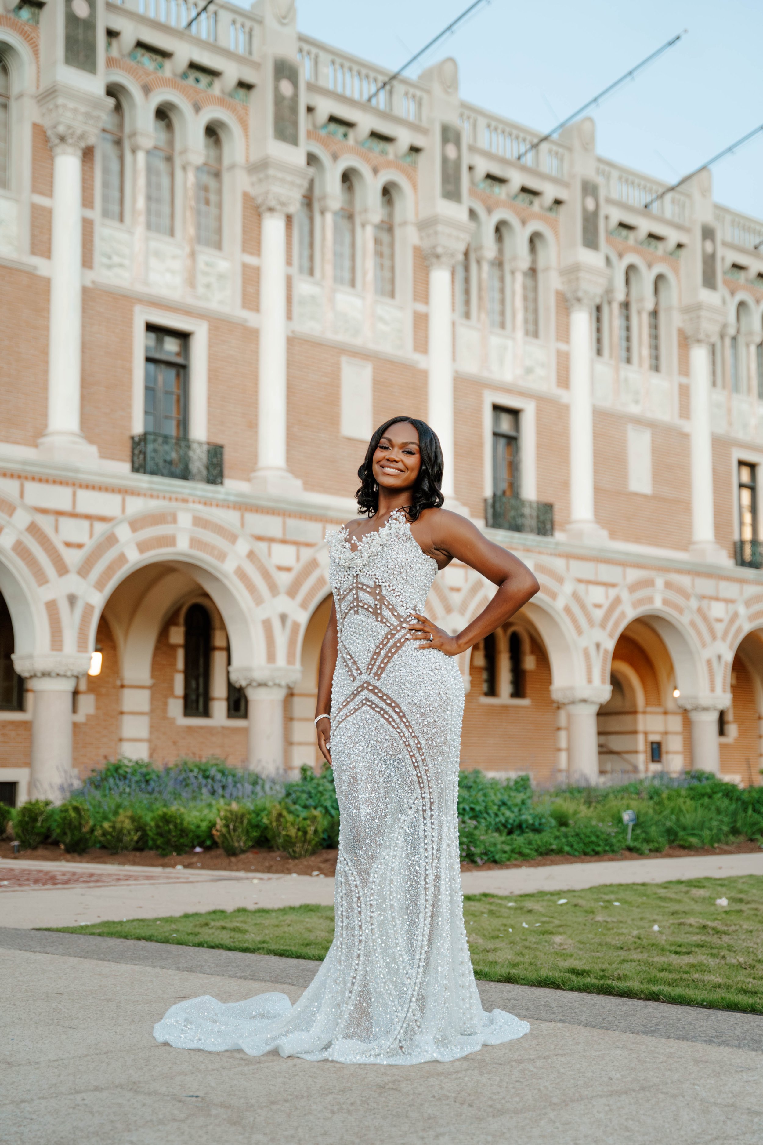 A woman in an elegant, beaded, silver evening gown stands in front of a historic brick and stone building with arched windows and columns, smiling and posing outdoors.