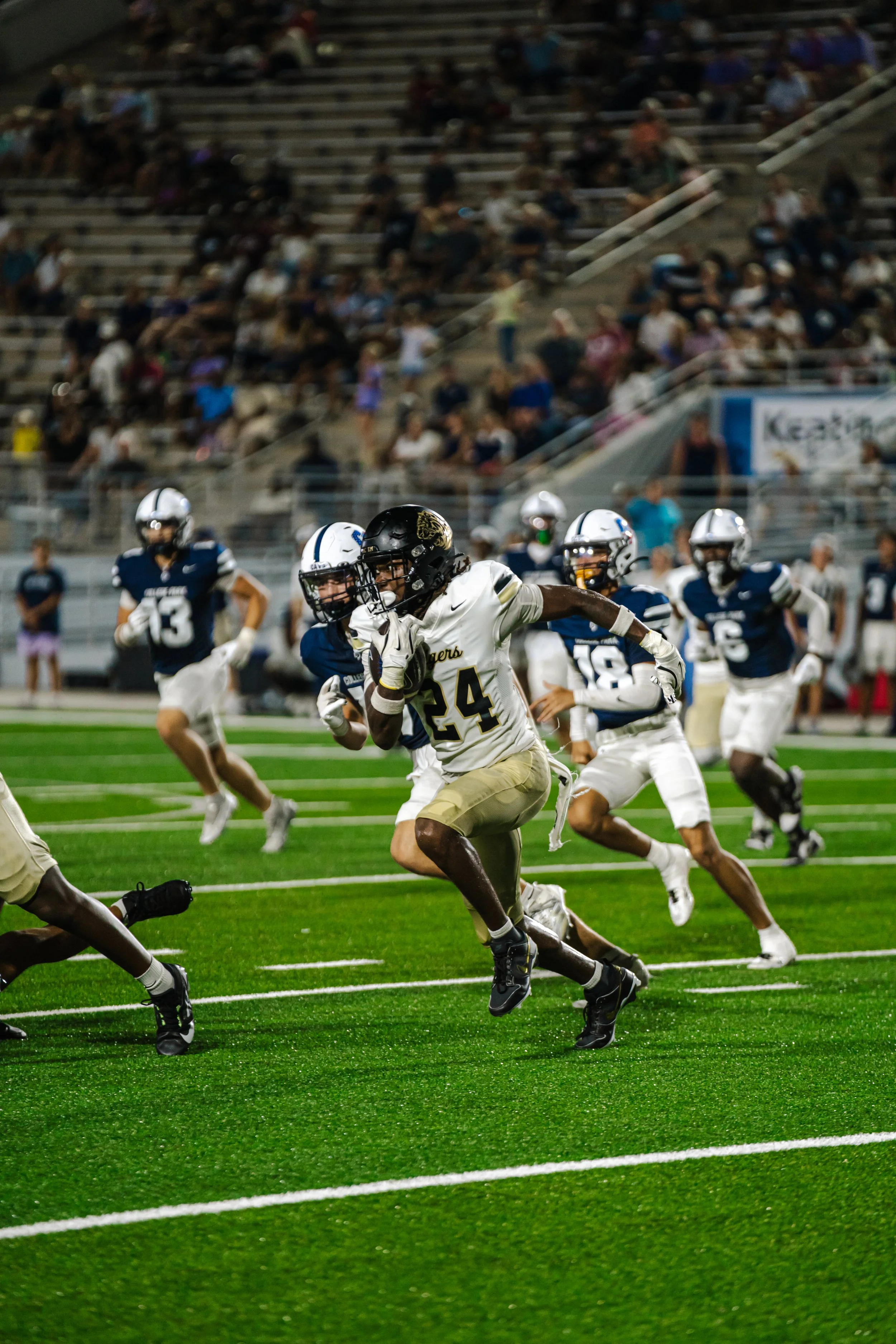 A football game at night, with players running on a green field, spectators sitting on bleachers, and stadium lights illuminating the scene.