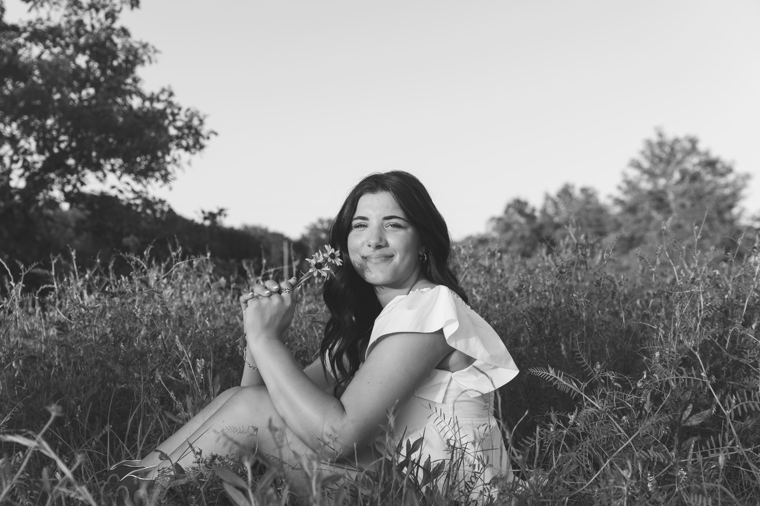 A woman with dark hair sitting in a field of tall grass holding a flower, smiling with her eyes closed, in black and white.