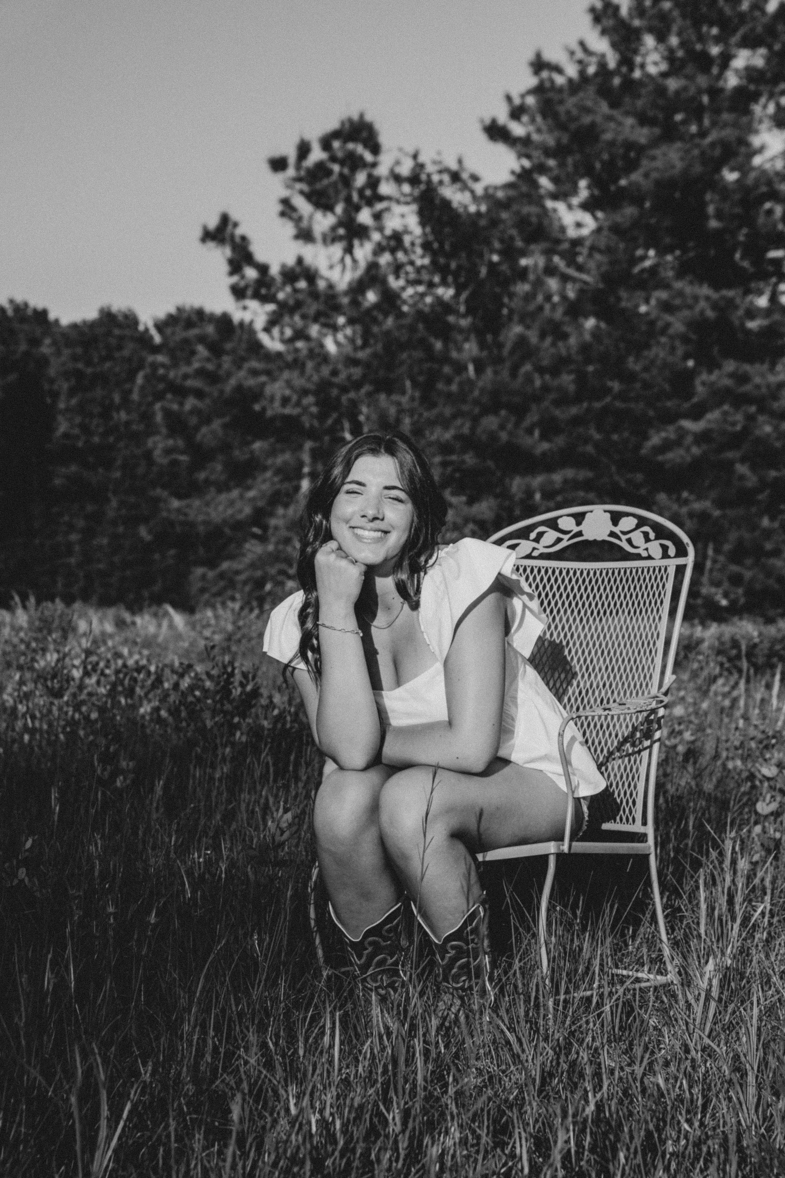 A young woman with long dark hair, smiling and sitting on a decorative metal chair outdoors in a grassy field with trees in the background. She is wearing a short-sleeved top and boots.