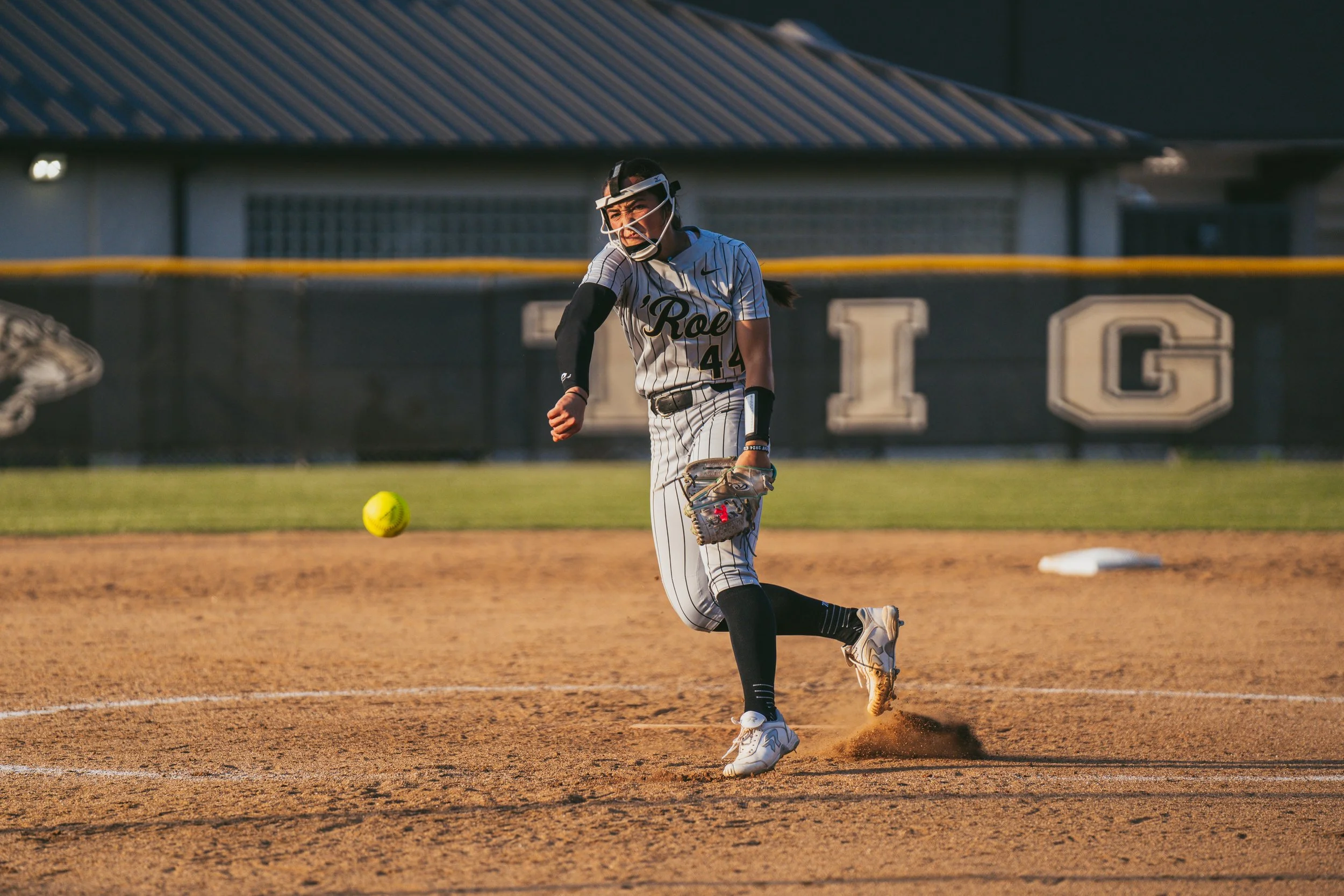 A female softball player in a white and black striped uniform with 'Roe' on the chest, number 44, is fielding a ball on her glove during a game on a dirt field, with her hair in a ponytail, wearing protective face gear, and the background shows a fen