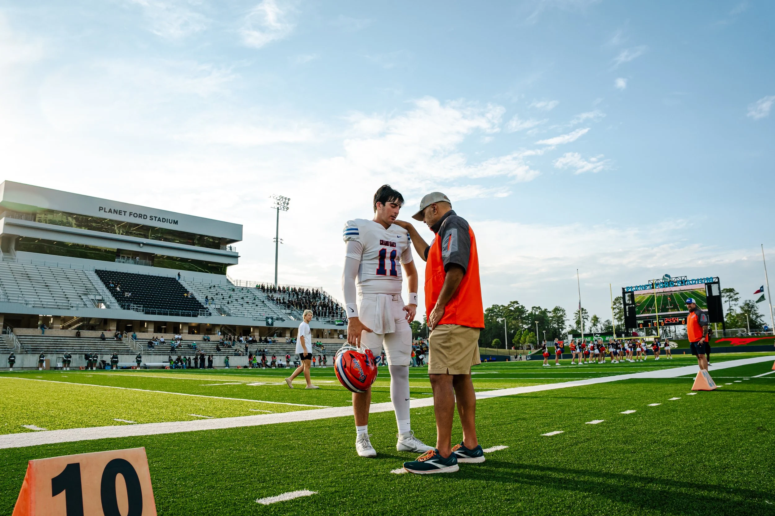 A football player in uniform standing on the field talking to a man in a gray hoodie and orange vest at a stadium, with other players and spectators visible in the background during late afternoon or early evening.