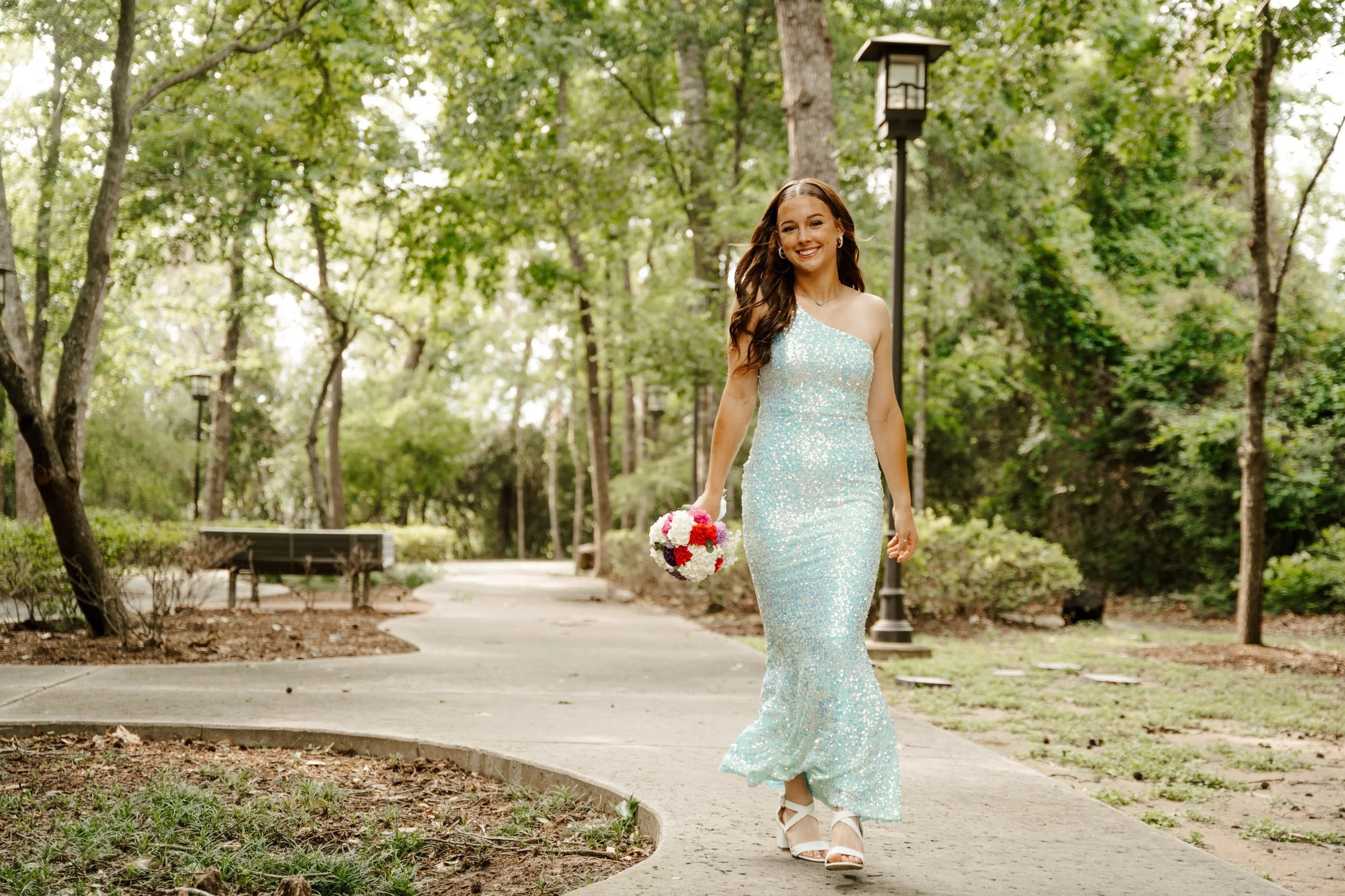A woman in a shimmering blue dress walking on a park pathway, holding a bouquet of flowers, surrounded by trees and greenery.