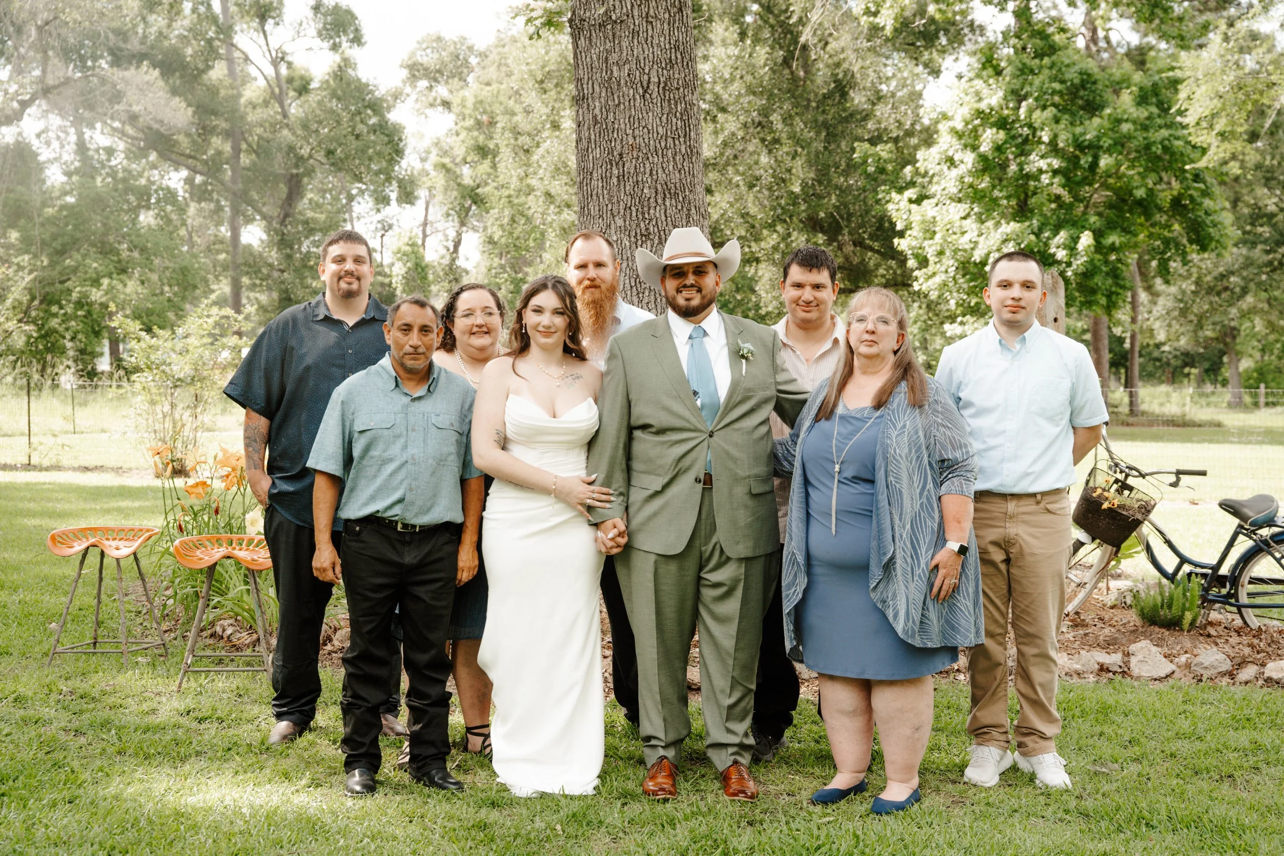 Group of nine diverse people standing outdoors on a lawn with trees in the background, some sitting on orange chairs, celebrating a wedding or special occasion.