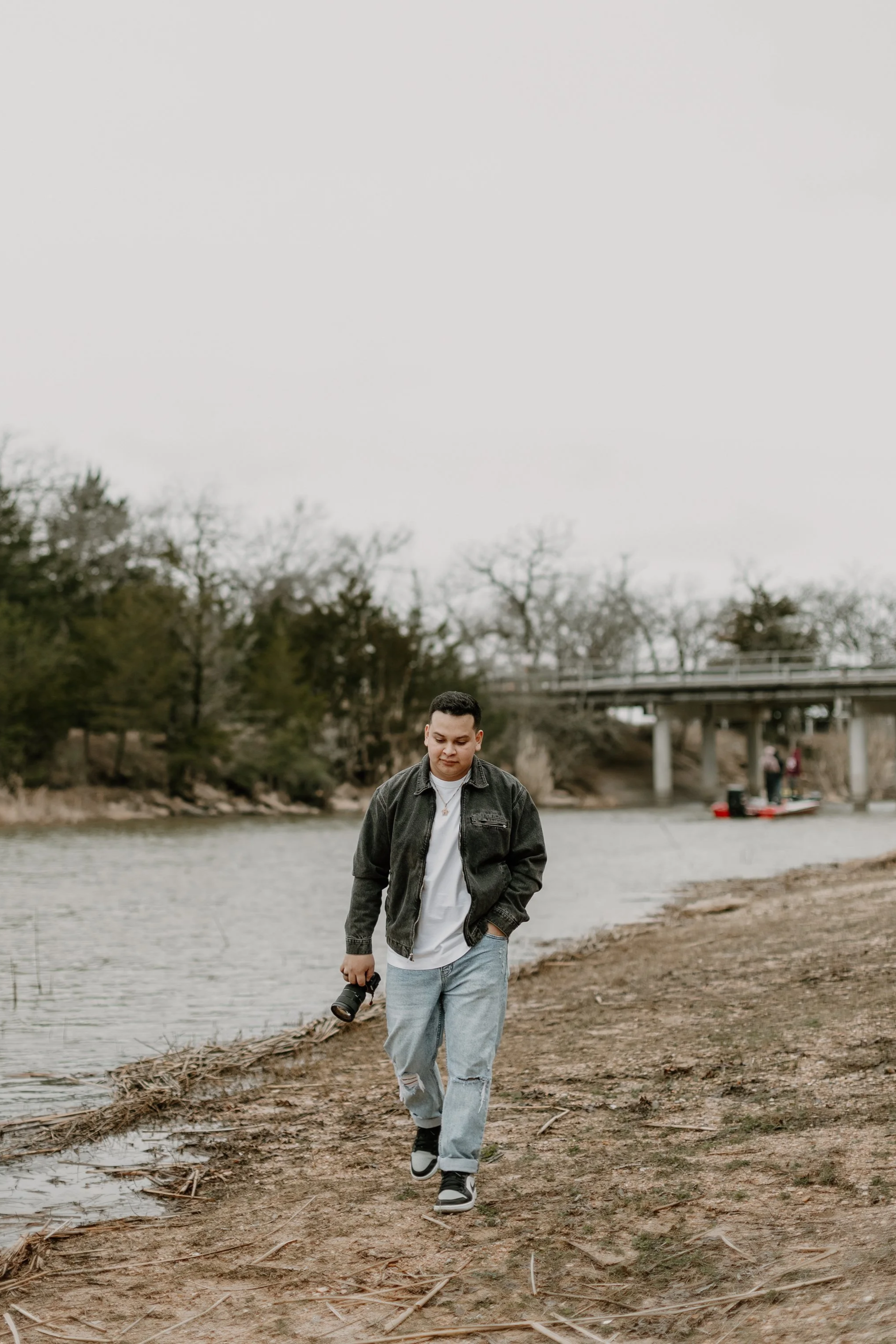 A young man walking along a muddy riverbank while holding a camera in his right hand, wearing a black jacket, white t-shirt, ripped jeans, and black sneakers with white soles, under an overcast sky, with trees and a bridge in the background.