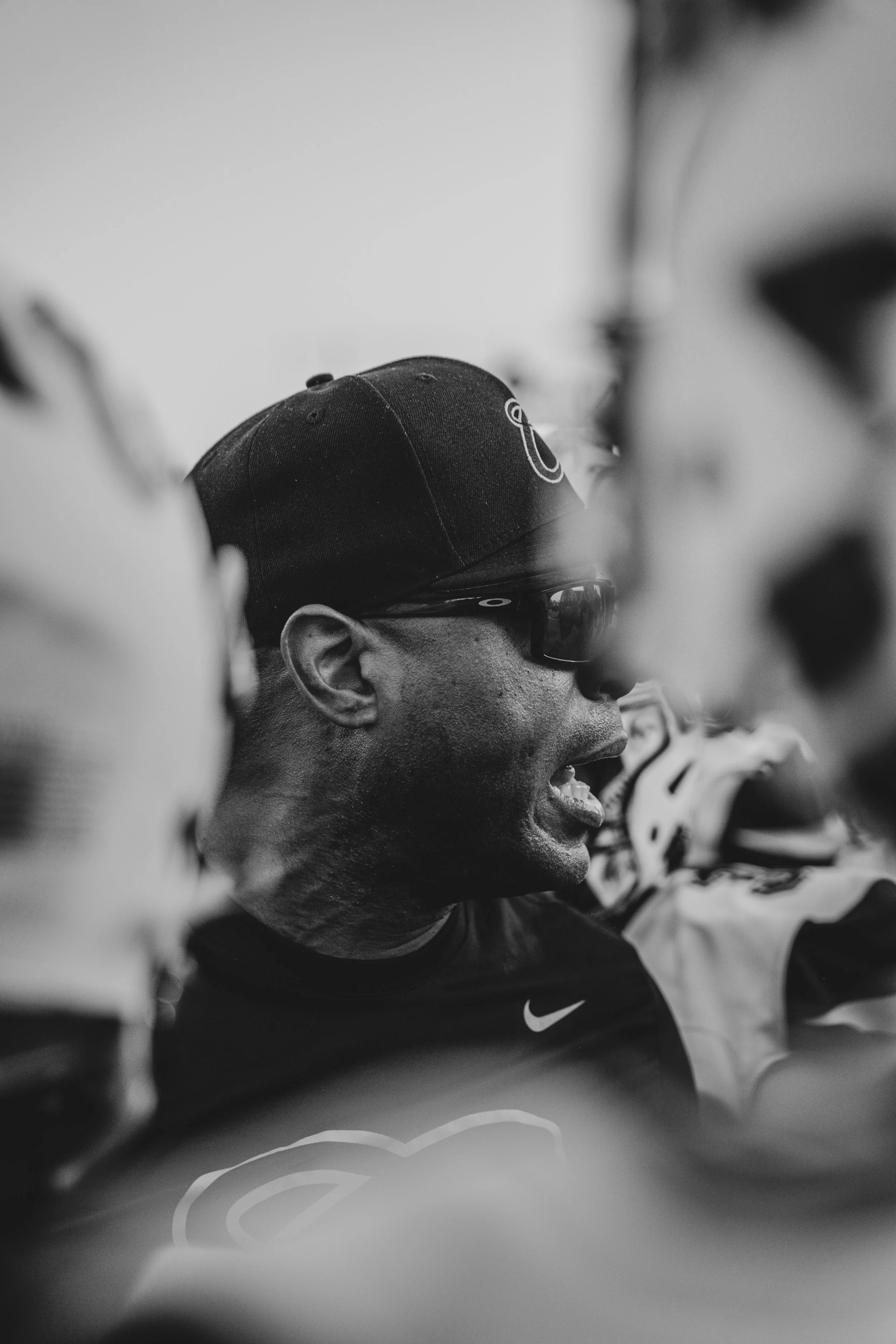 Black and white photo of a cyclist's profile, wearing a cap and sunglasses, surrounded by other cyclists in a race or group ride.