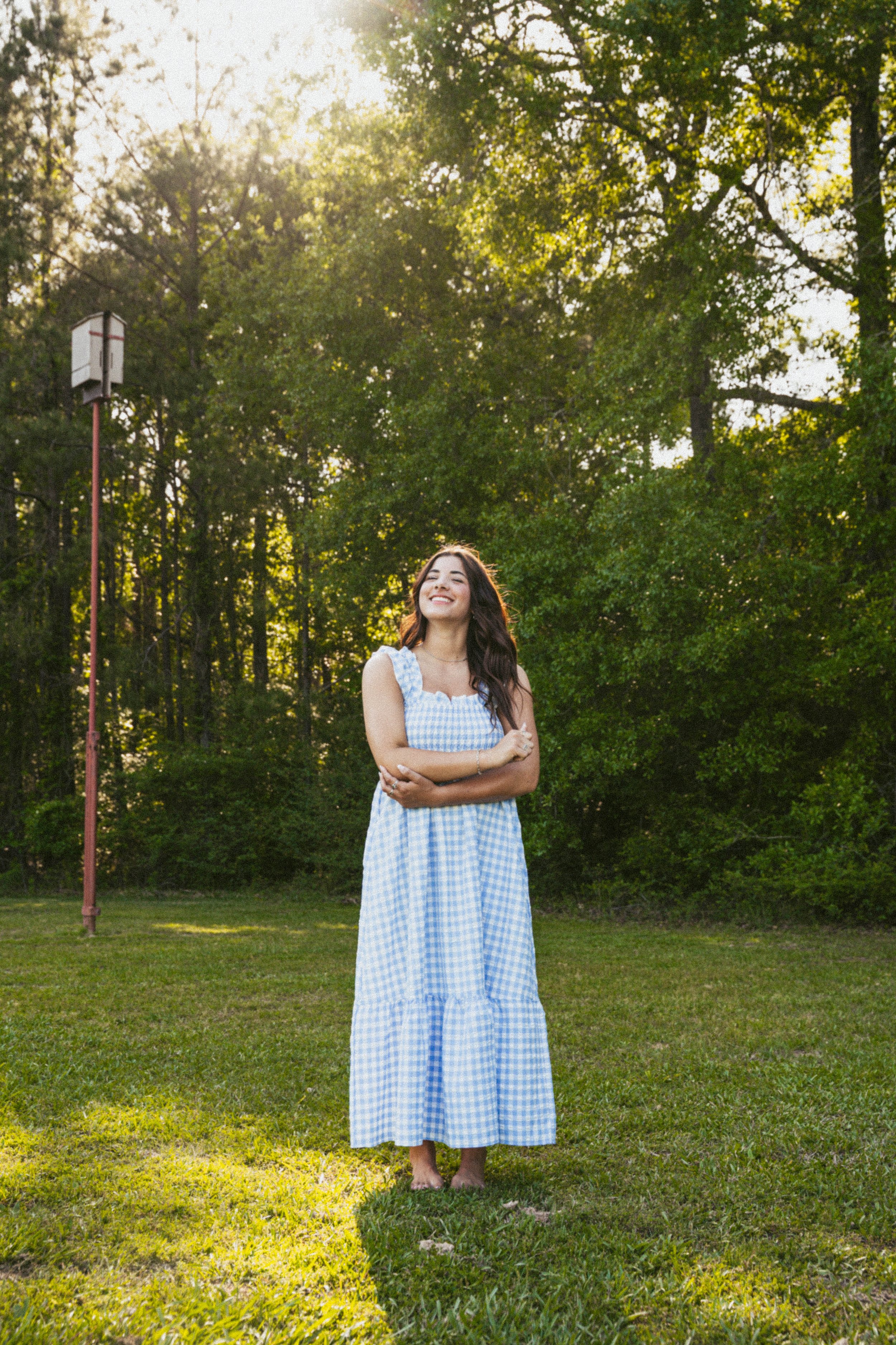 A young woman with long dark hair standing outdoors in a grassy area, smiling with her eyes closed and arms crossed, wearing a light blue and white checkered sleeveless dress, with tall trees and a birdhouse on a pole in the background during late af