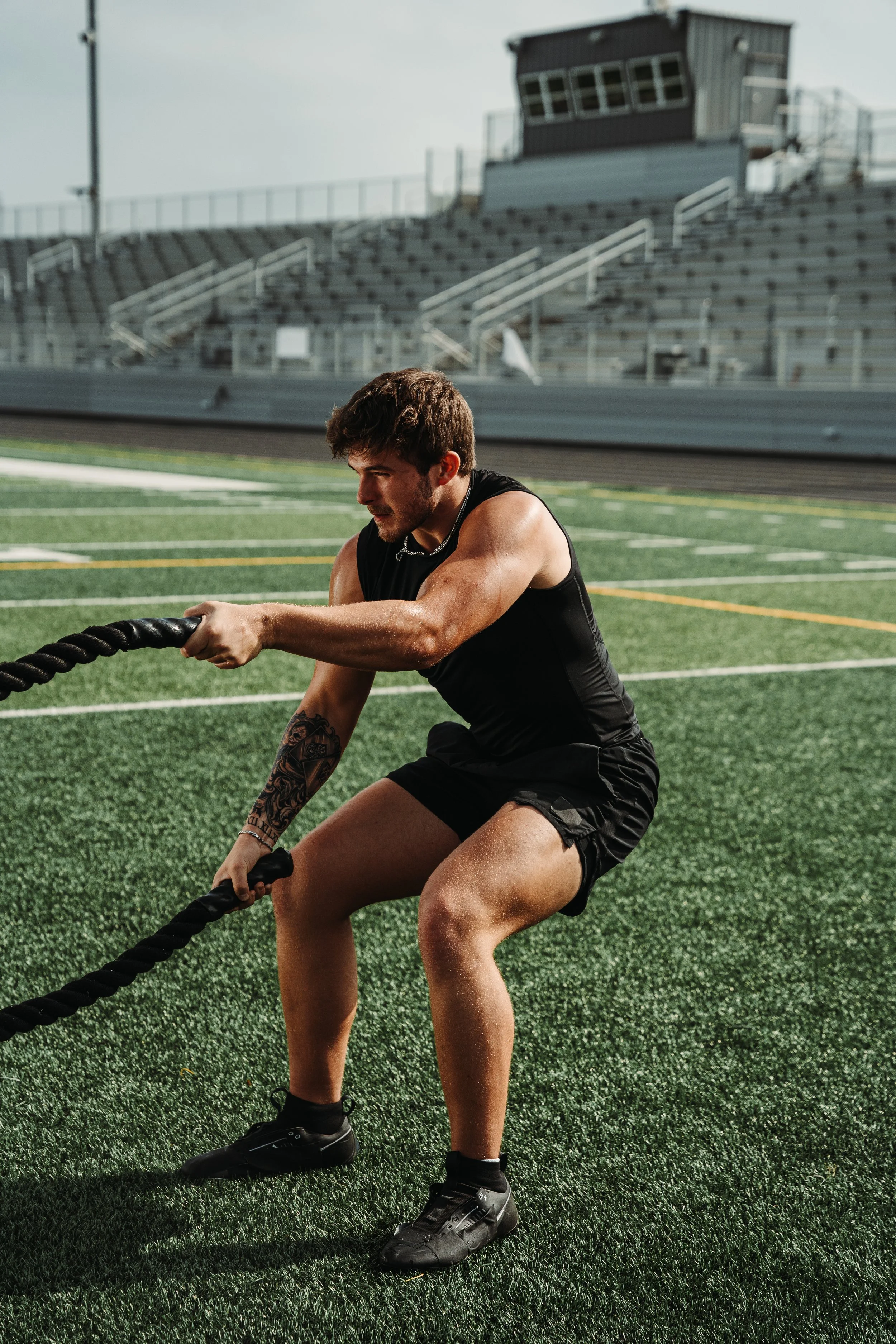 A young man with a tattoo on his right arm is working out with battle ropes on a green sports field. He is wearing a black sleeveless shirt, shorts, and black athletic shoes. Bleachers and a building can be seen in the background.