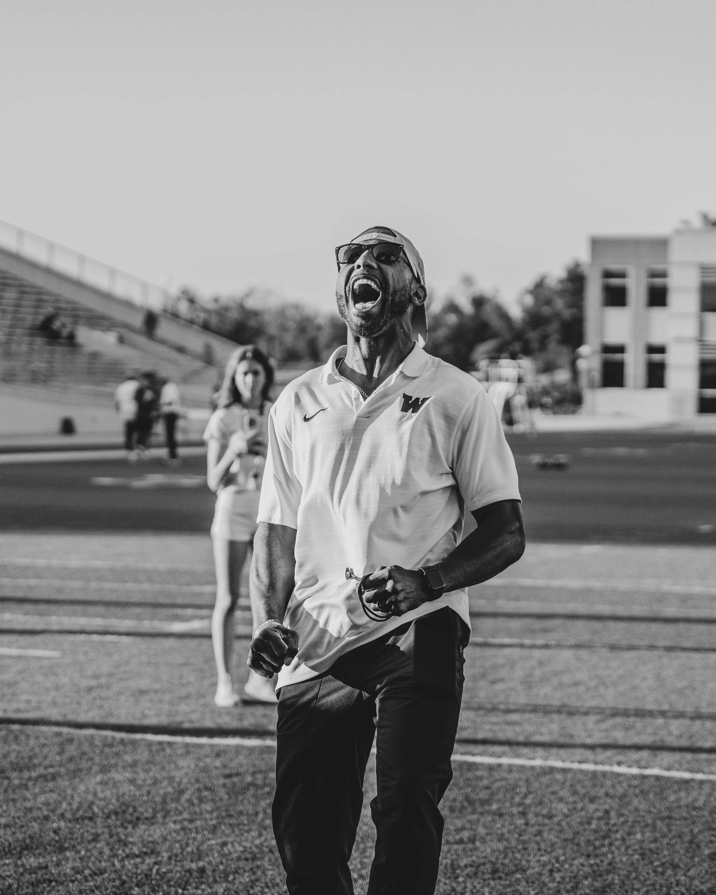 A Black man with a beard, wearing sunglasses, a cap backwards, and a Nike shirt, running on a track with an intense expression, mouth open, in black and white.