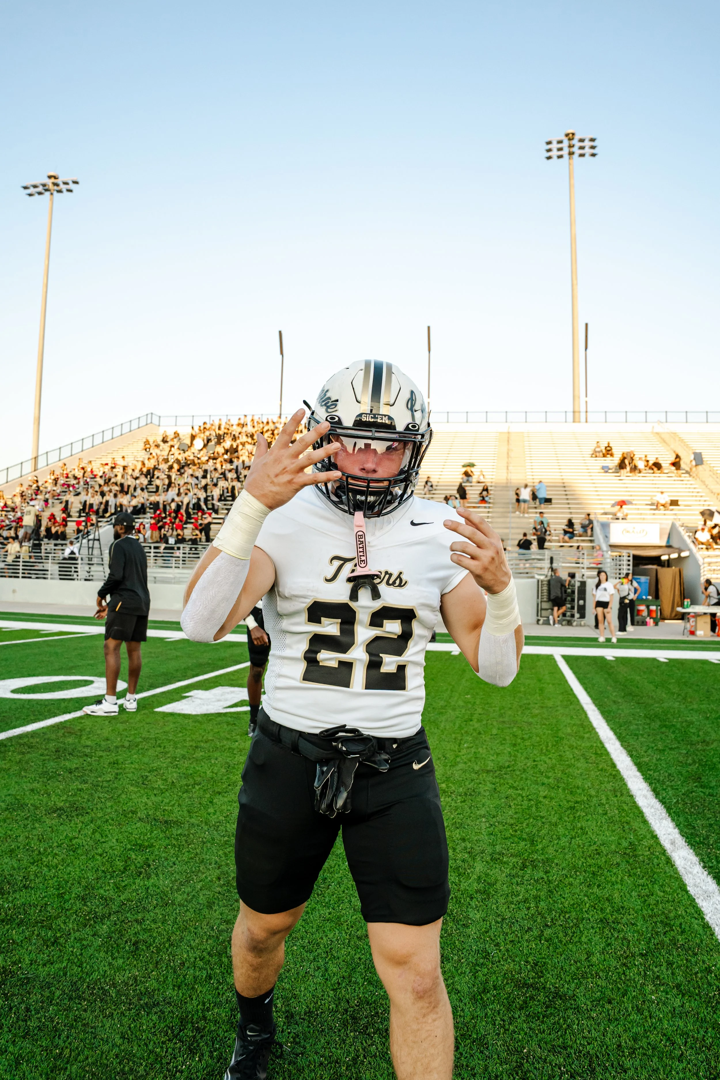 A football player wearing a white jersey with the number 22 and a black helmet, standing on a football field with his hand near his face mask. The background shows spectators in the stands and stadium lights.