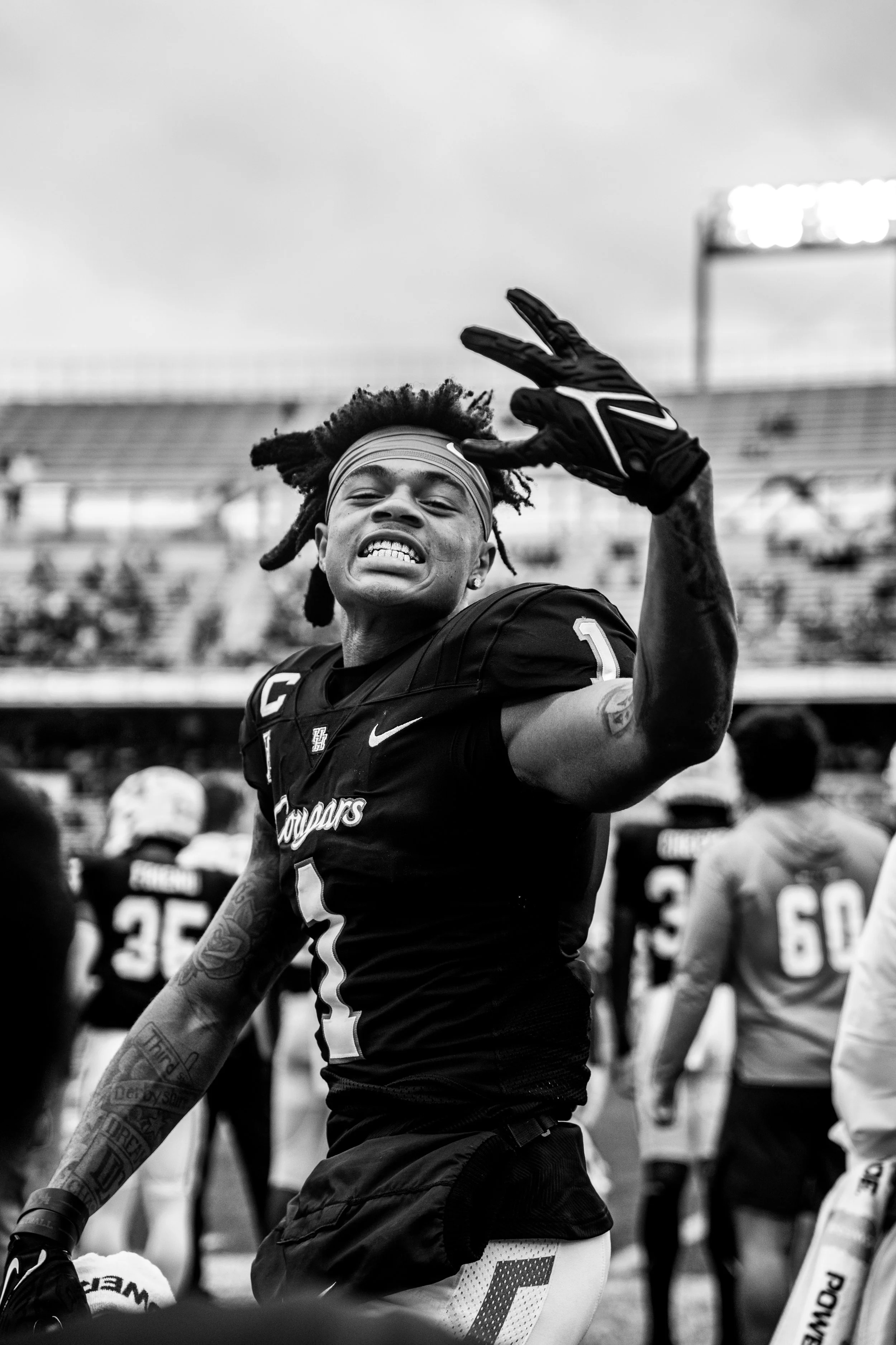 Close-up of an African American football player with dreadlocks and a headband, wearing a jersey with the word 'Cougars' and the number 1, smiling and waving with one hand covered in a glove, on a football field with teammates in the background.