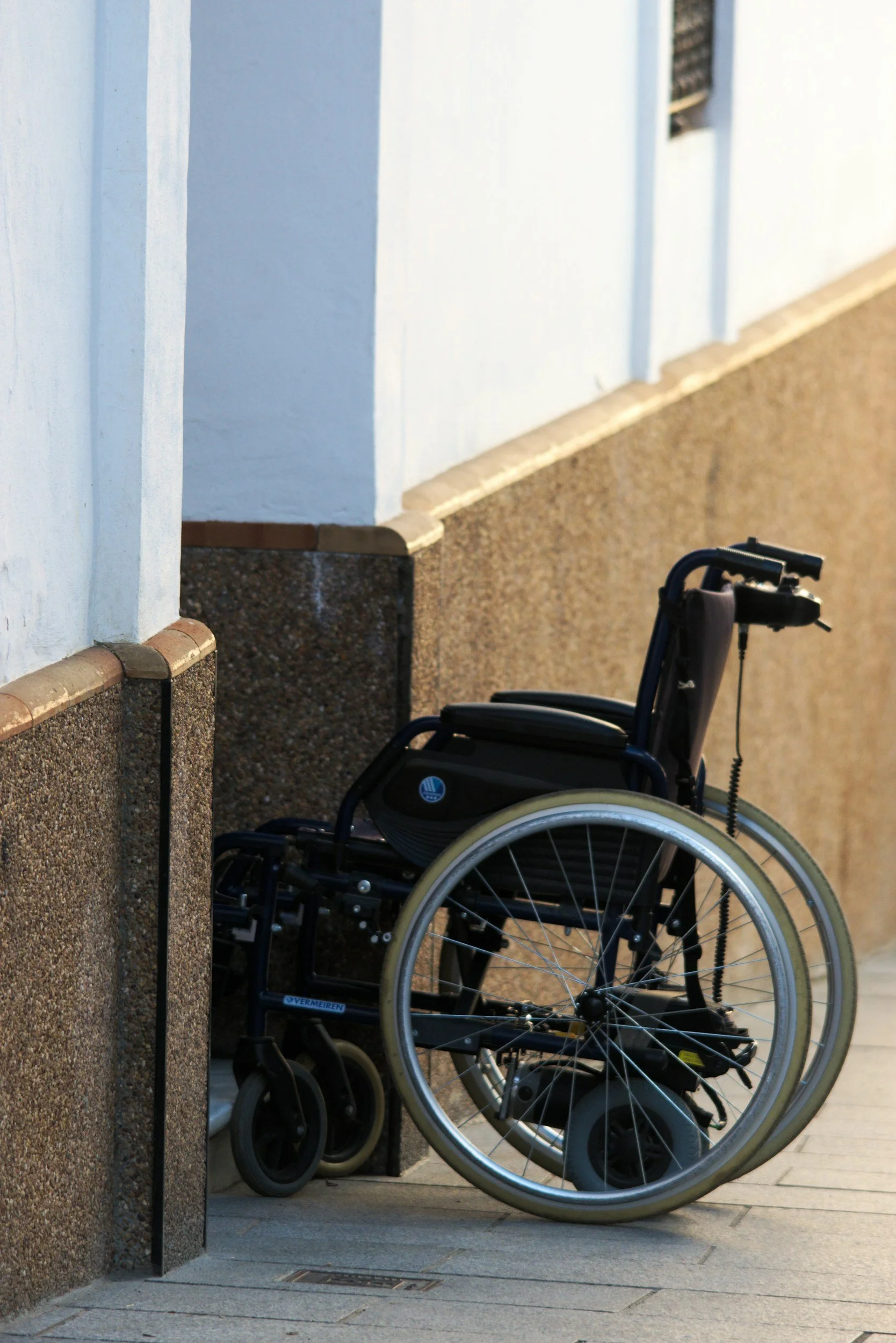 A wheelchair parked next to a building wall with brown and beige textured bricks on the lower part and plain white painted surface on the upper part.