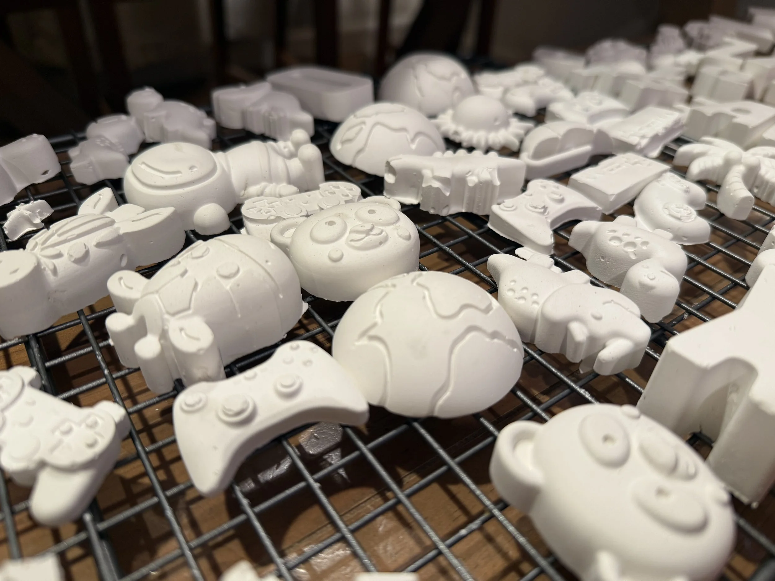 White ceramic figurines of various playful characters, such as animals, robots, and cartoon faces, arranged on a metal cooling rack.