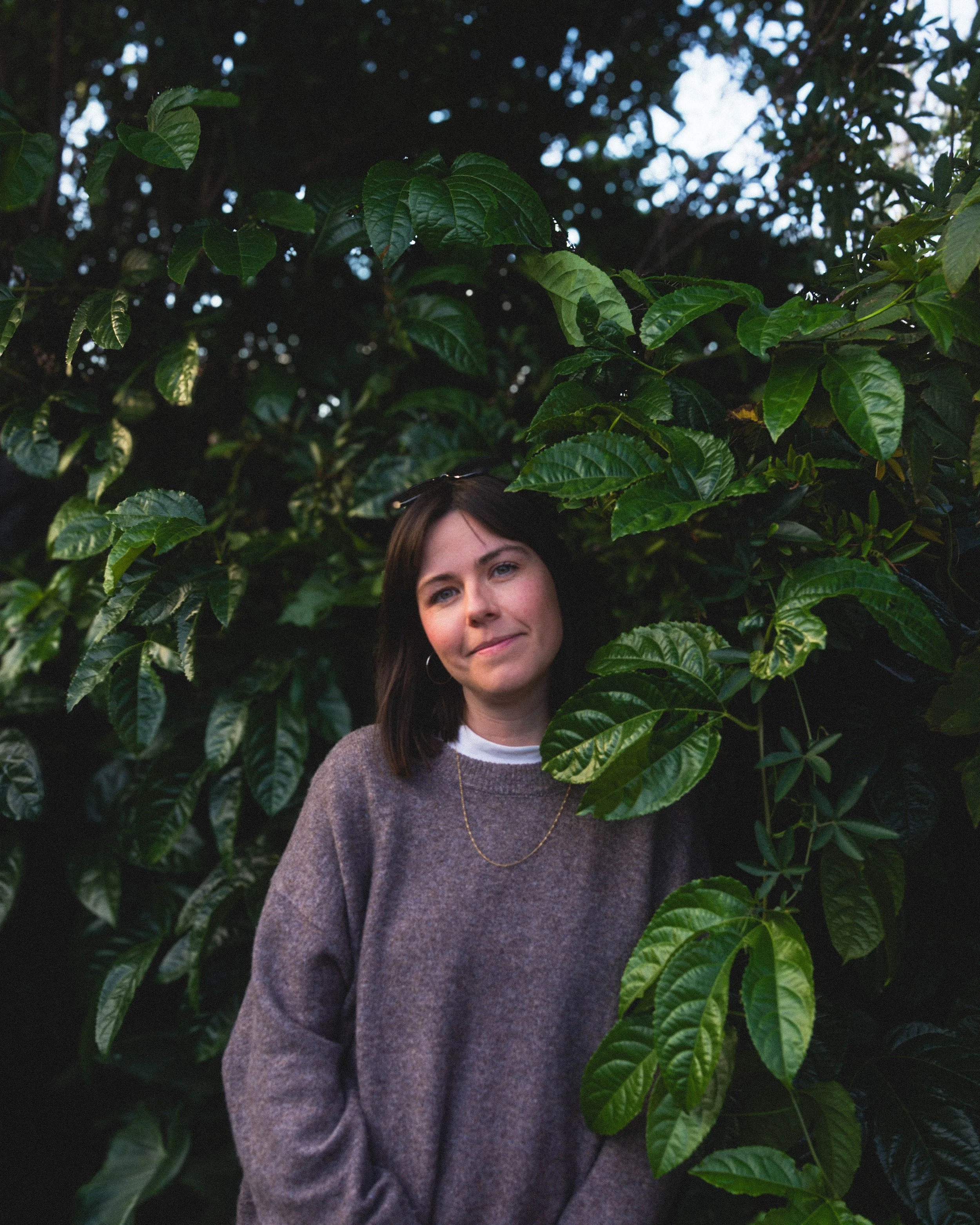 A woman with brown hair wearing a gray sweater and a gold necklace standing among dense green foliage with large dark green leaves.