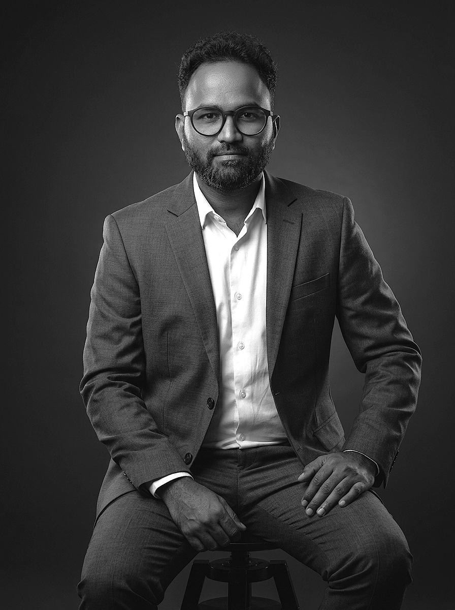 Black-and-white portrait of a man wearing glasses, a white shirt, and a suit, sitting on a stool with a dark background.