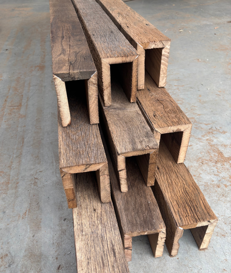 Stacked wooden planks with a U-shaped profile, arranged in a pyramid shape on a concrete surface.