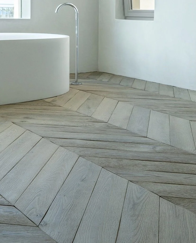 Modern bathroom with curved white bathtub, chrome floor-mounted faucet, white walls, and herringbone-patterned wooden floor.