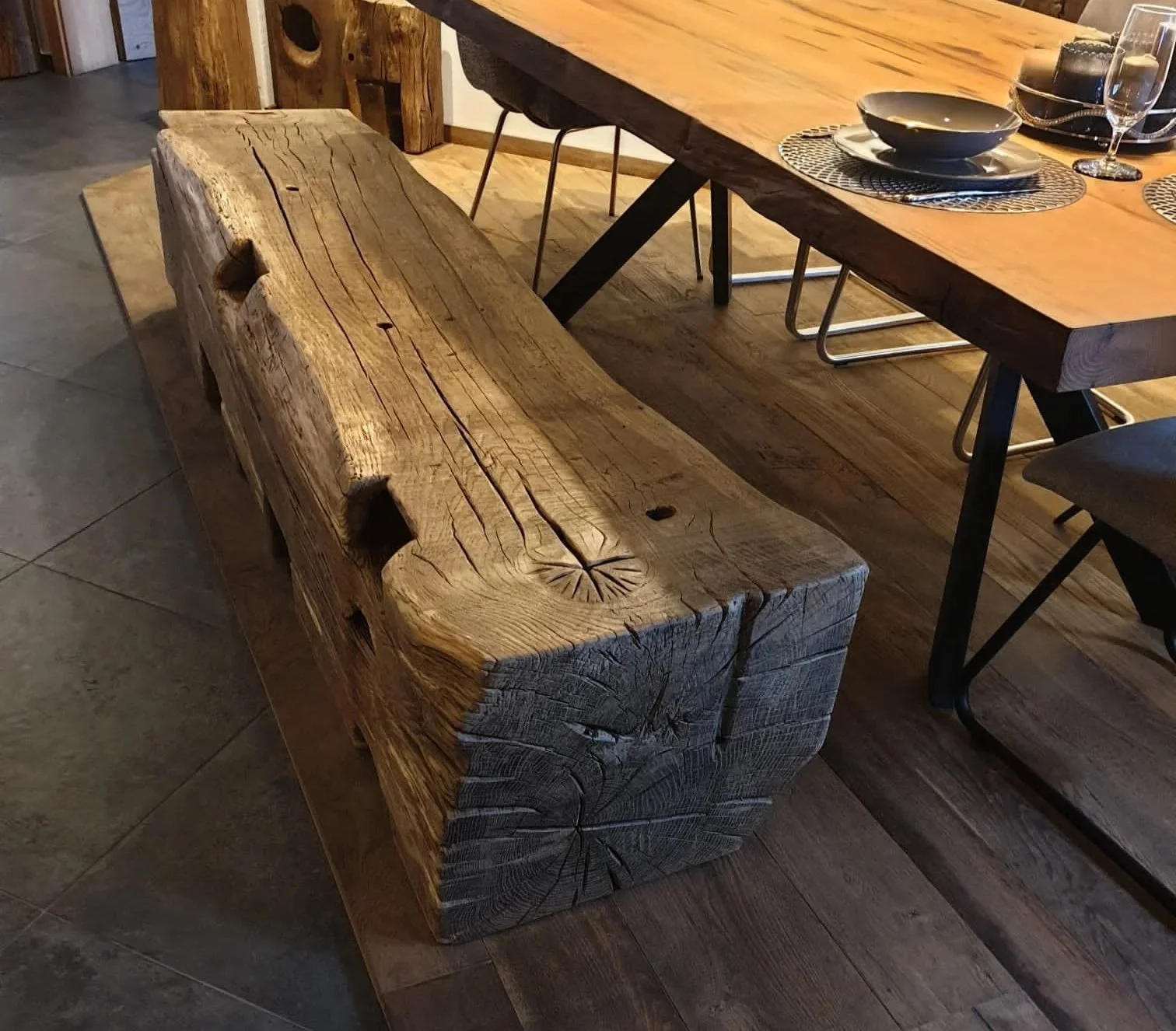A rustic wooden bench with a rough-hewn, textured surface and visible wood grain in a dining room, next to a polished wooden dining table.