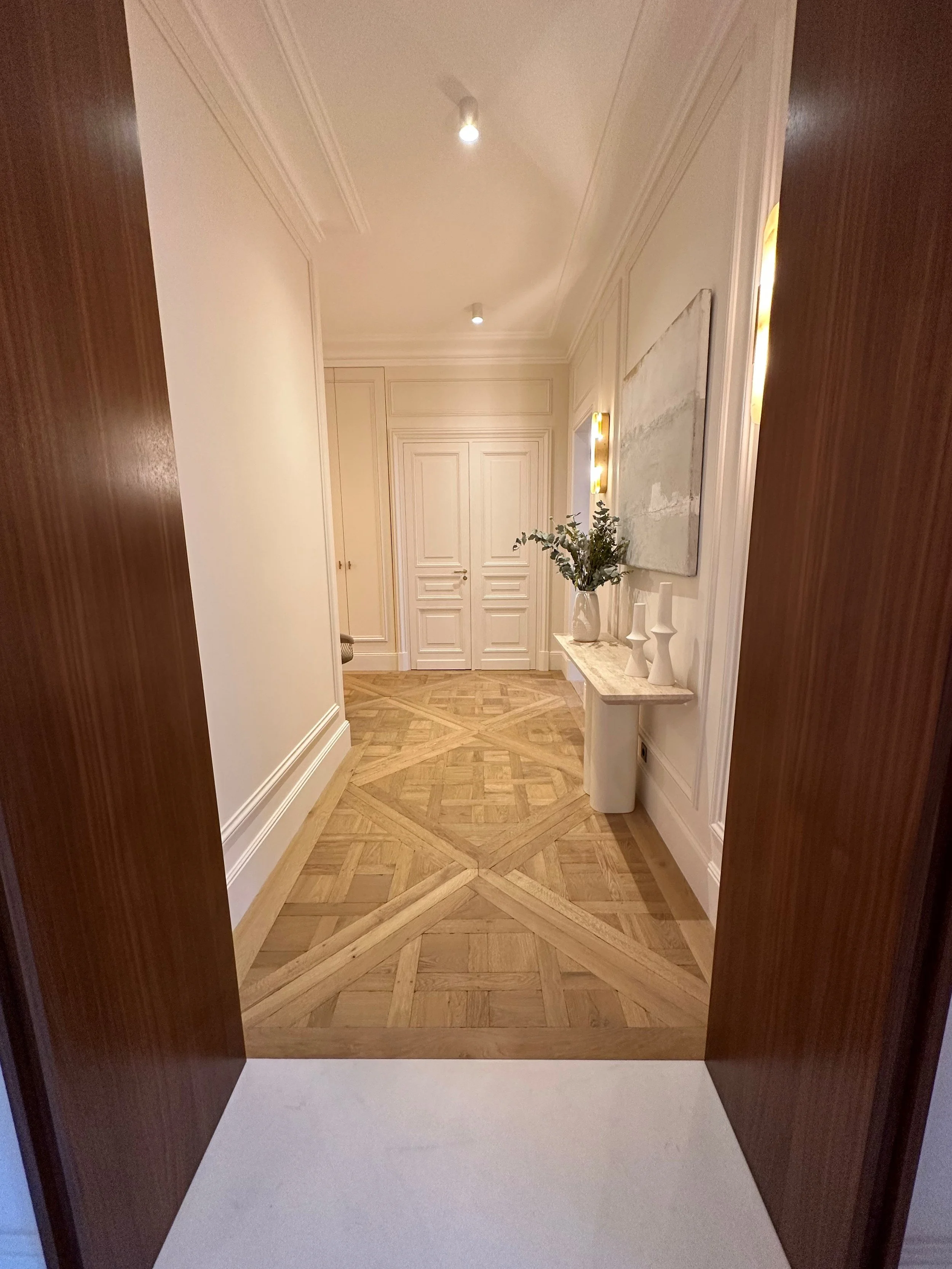 View from a room with dark wooden double doors, looking into a corridor with intricate parquet wood flooring, white walls, and elegant decor including a marble-topped table with vases and a large abstract painting.