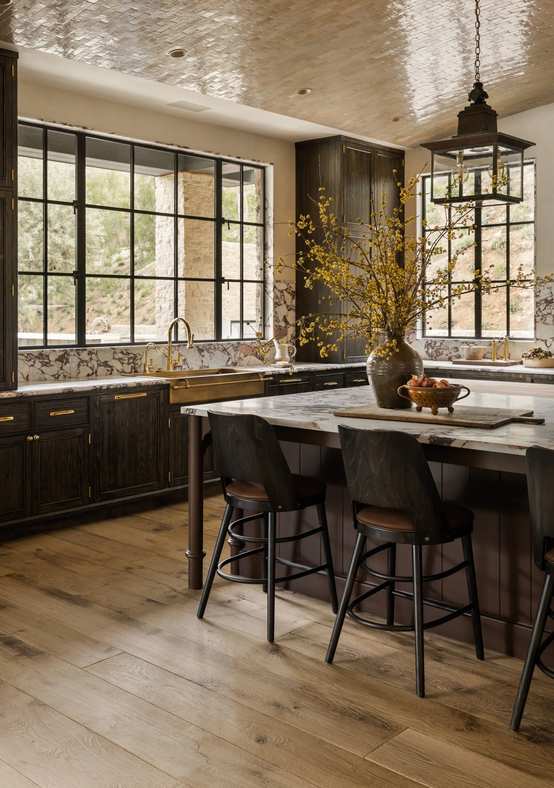 French oak flooring. Kitchen with dark wooden cabinets, marble countertops, large black grid windows, island bench and wooden chairs with black seats.