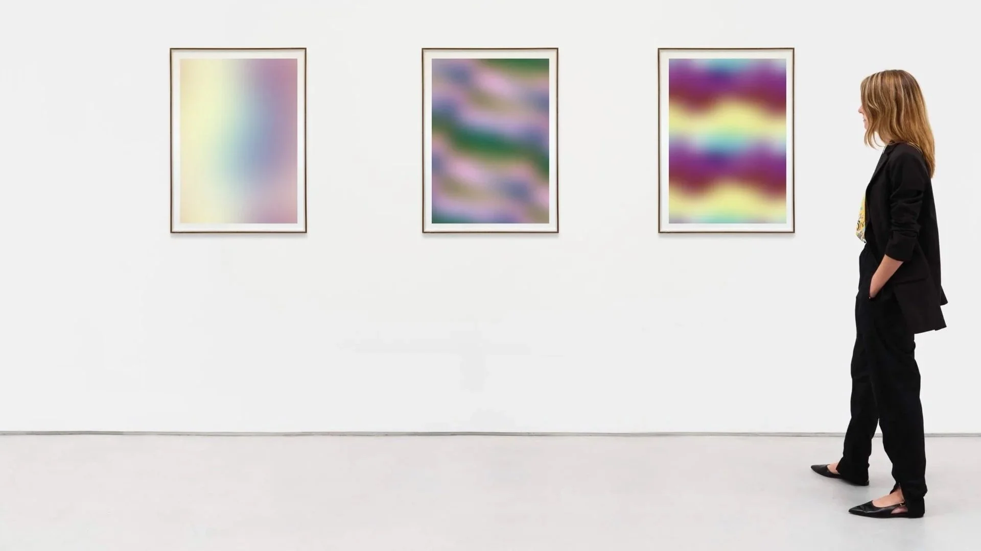 A woman dressed in black trousers and blazer standing in an art gallery, observing three framed abstract colorful paintings on a white wall.