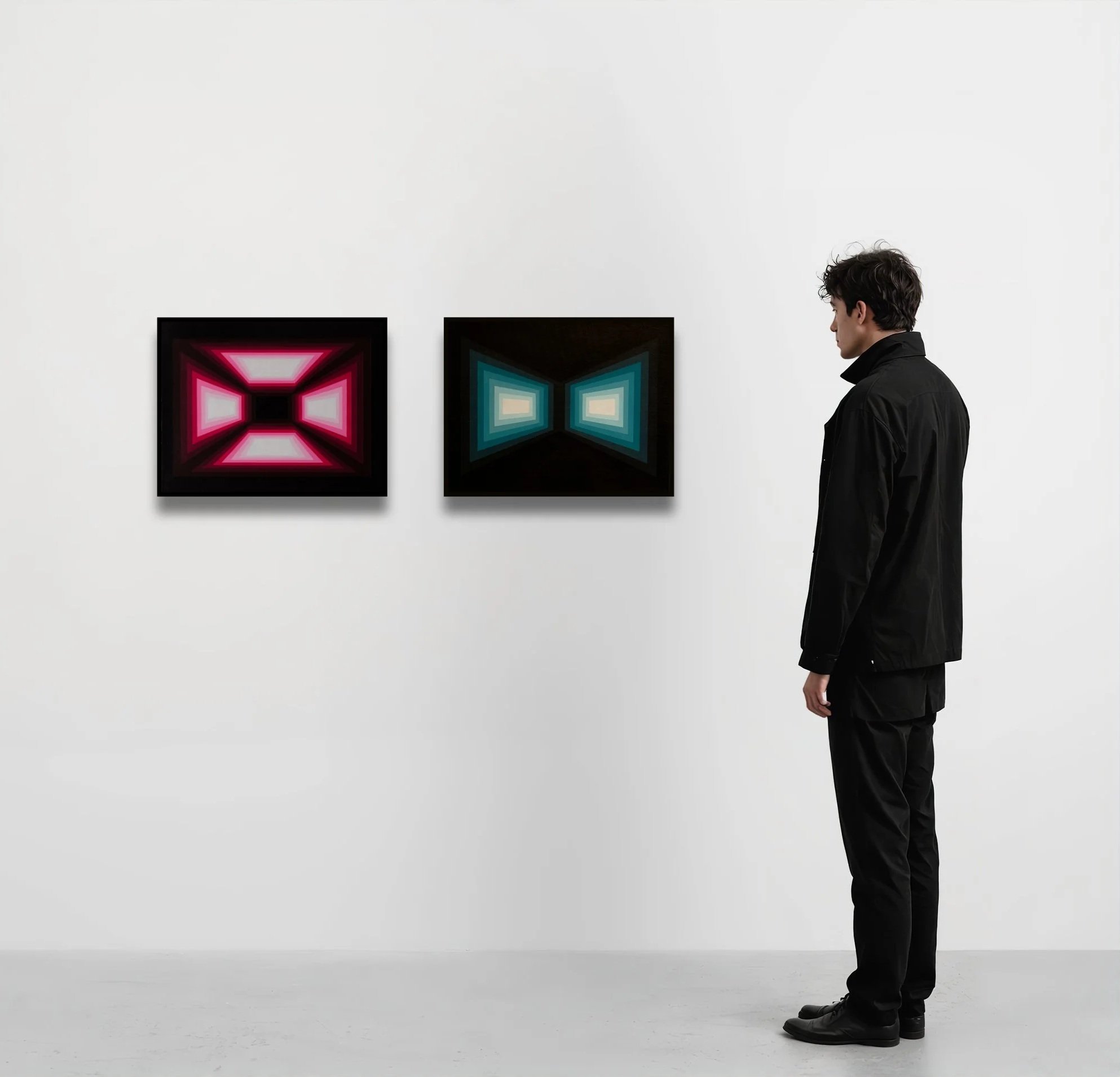A man with dark, curly hair dressed in a black jacket and pants stands in an art gallery next to two framed abstract digital artwork pieces. One has a red geometric design, the other blue, both with concentric shapes, against a white wall.