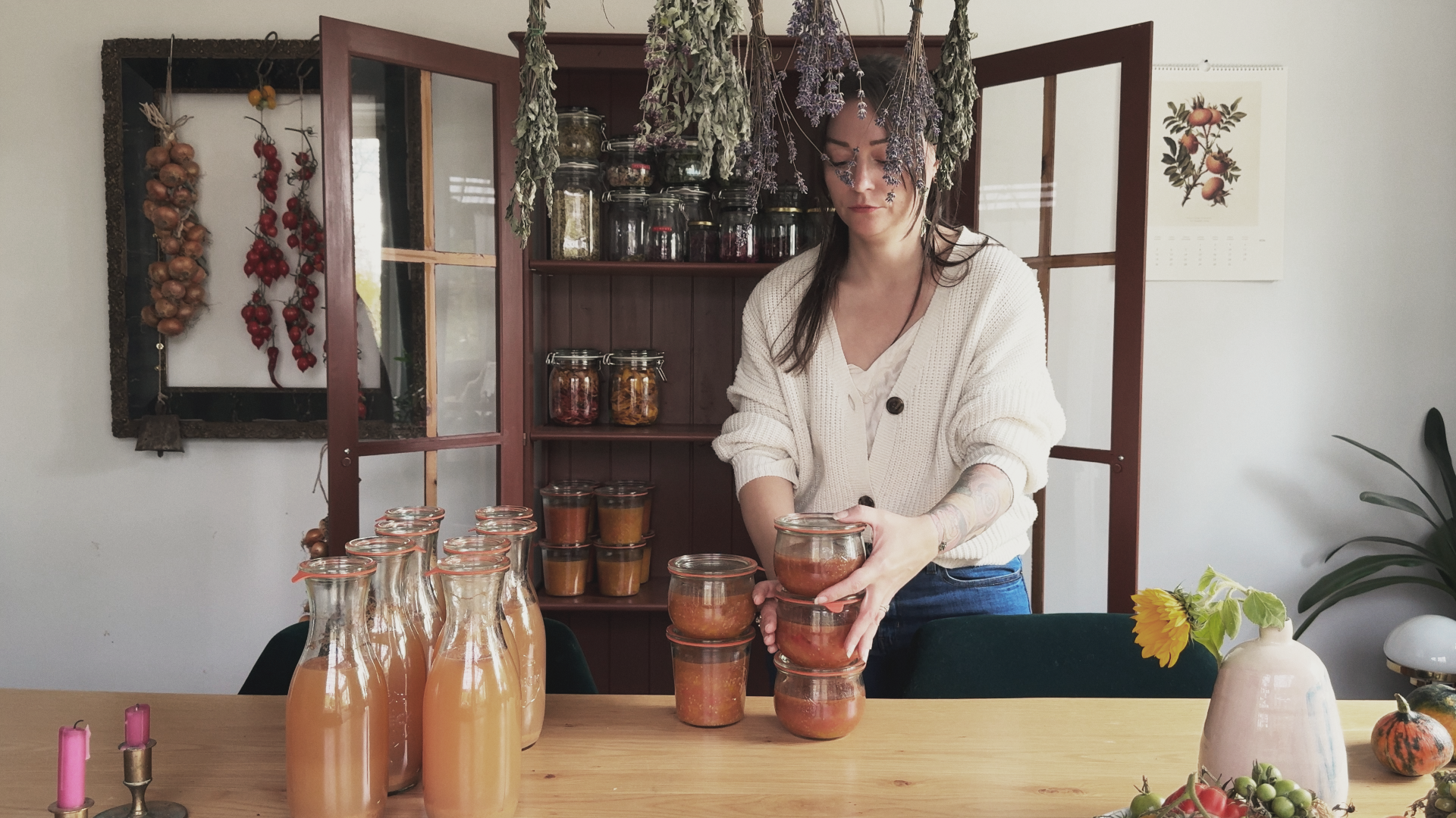 Stacking jars of homemade tomato sauce beside bottles of apple juice, with dried herbs hanging overhead